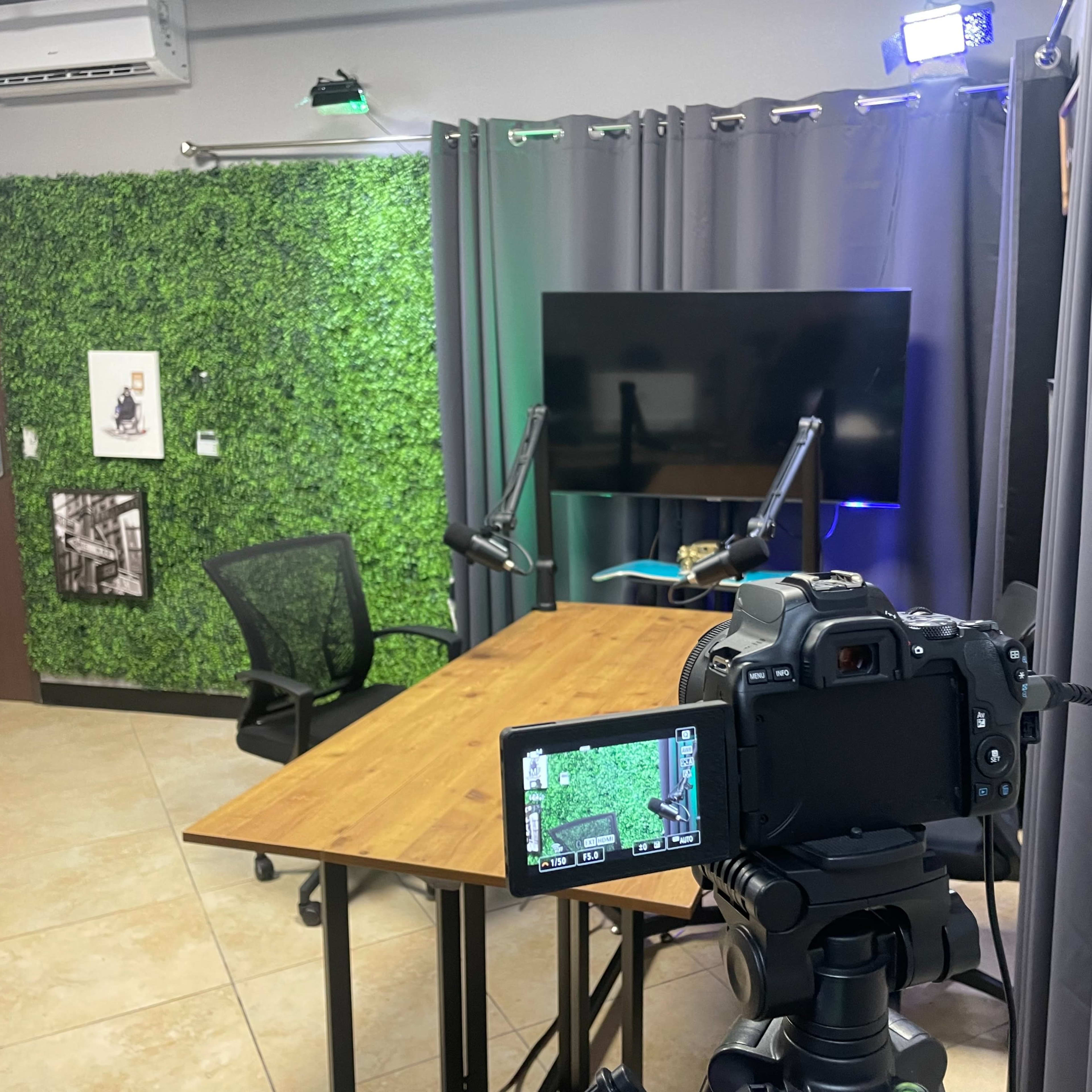 A video recording setup with a camera on a tripod focused on a wooden table, surrounded by green foliage, curtains, and a large screen.