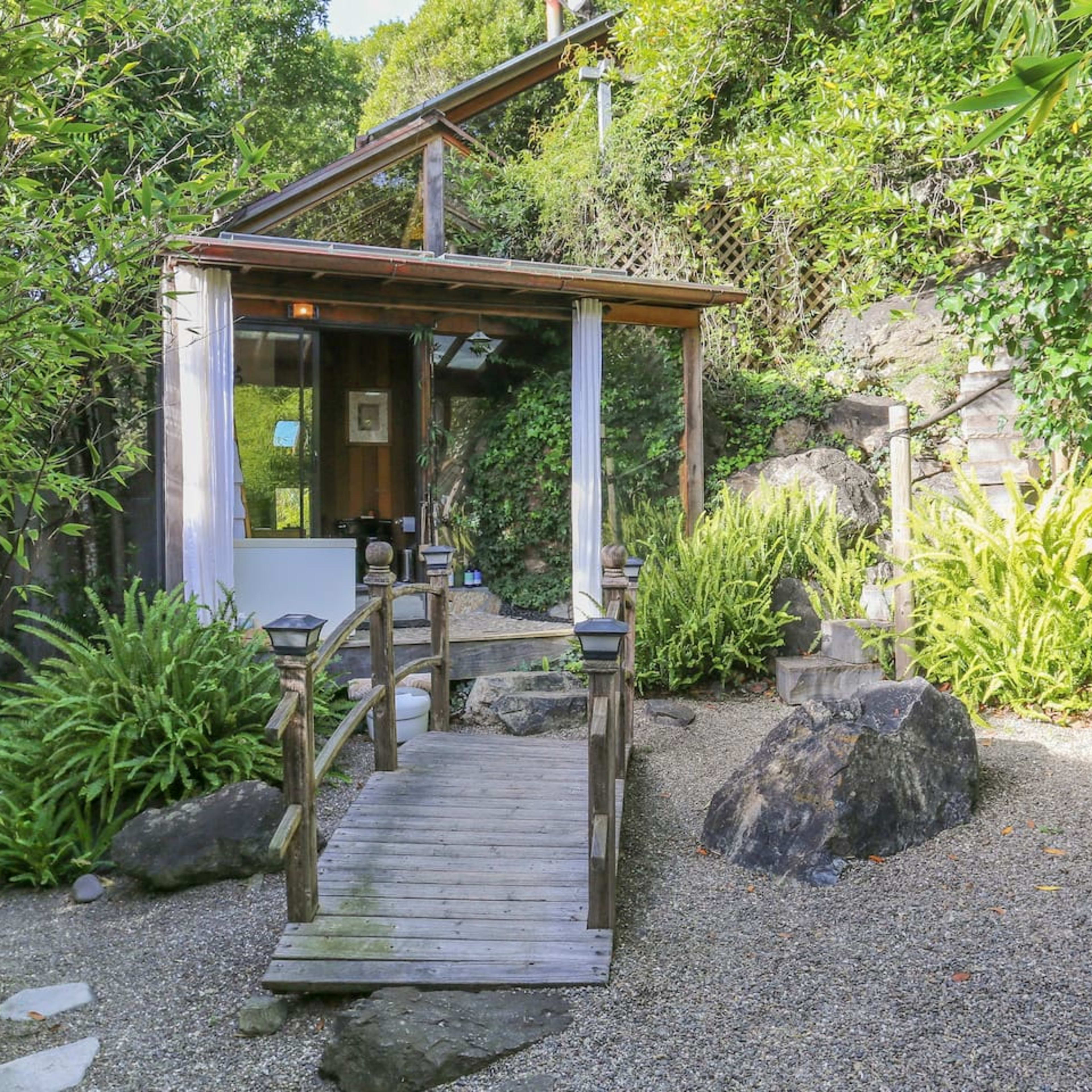 A wooden bridge leads to a small building surrounded by rocks and lush greenery in a landscaped garden.