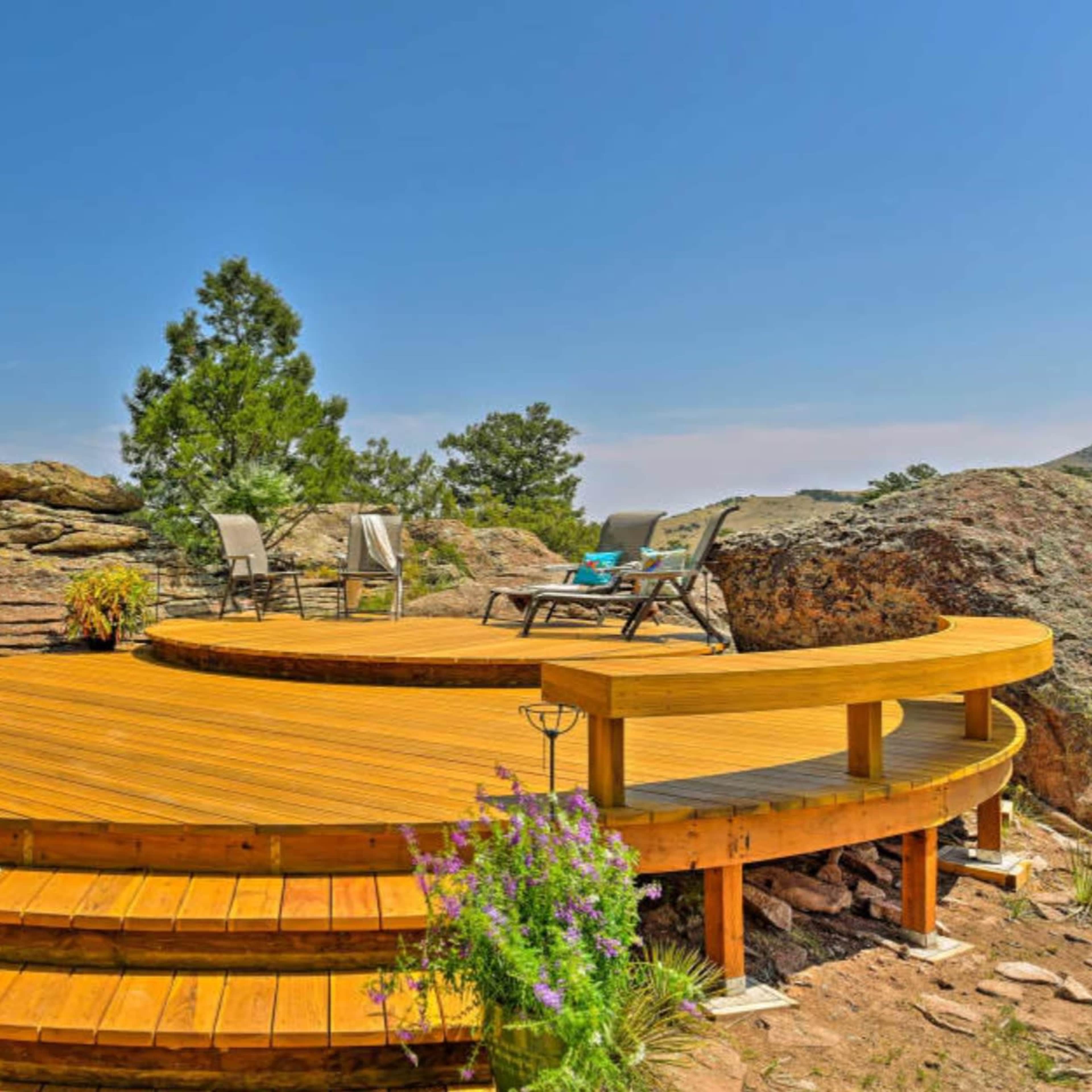 A circular wooden deck with lounge chairs, surrounded by large rocks and greenery under a clear blue sky.