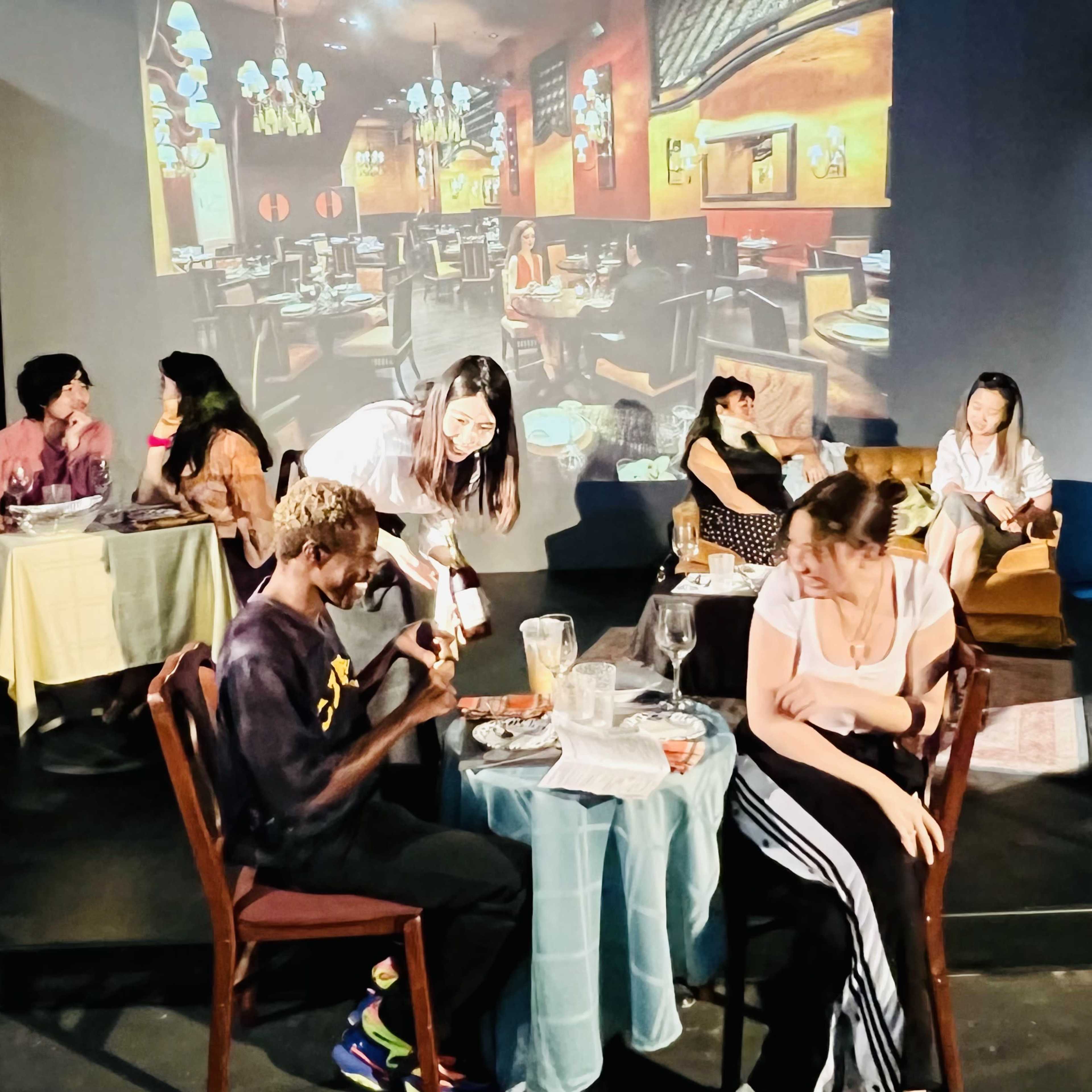A group of actors performs a scene in a dimly lit restaurant setting with projected images in the background.