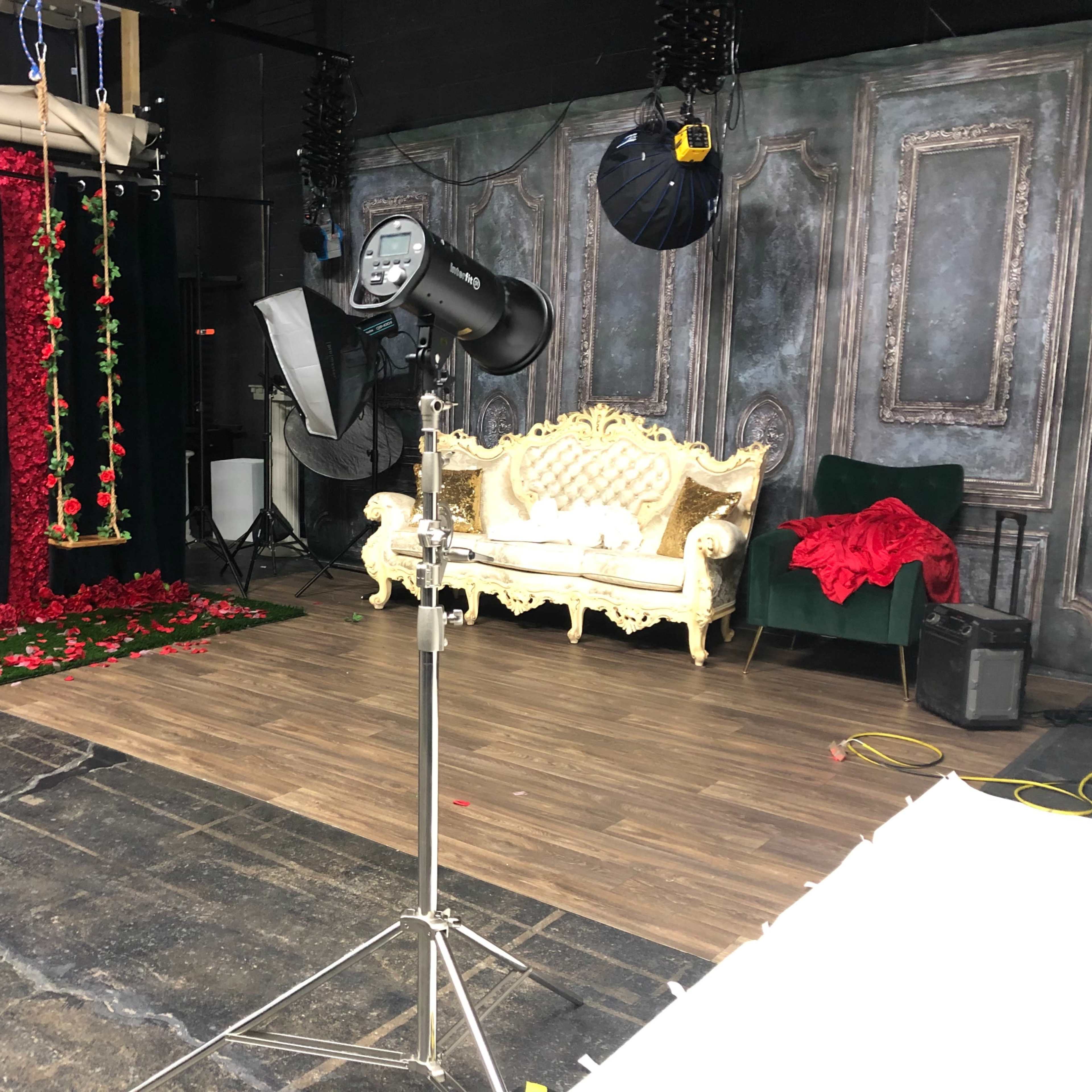 A studio setup with a decorative couch, a swing adorned with roses, and various lighting equipment against a backdrop of draped fabric and a wooden floor.