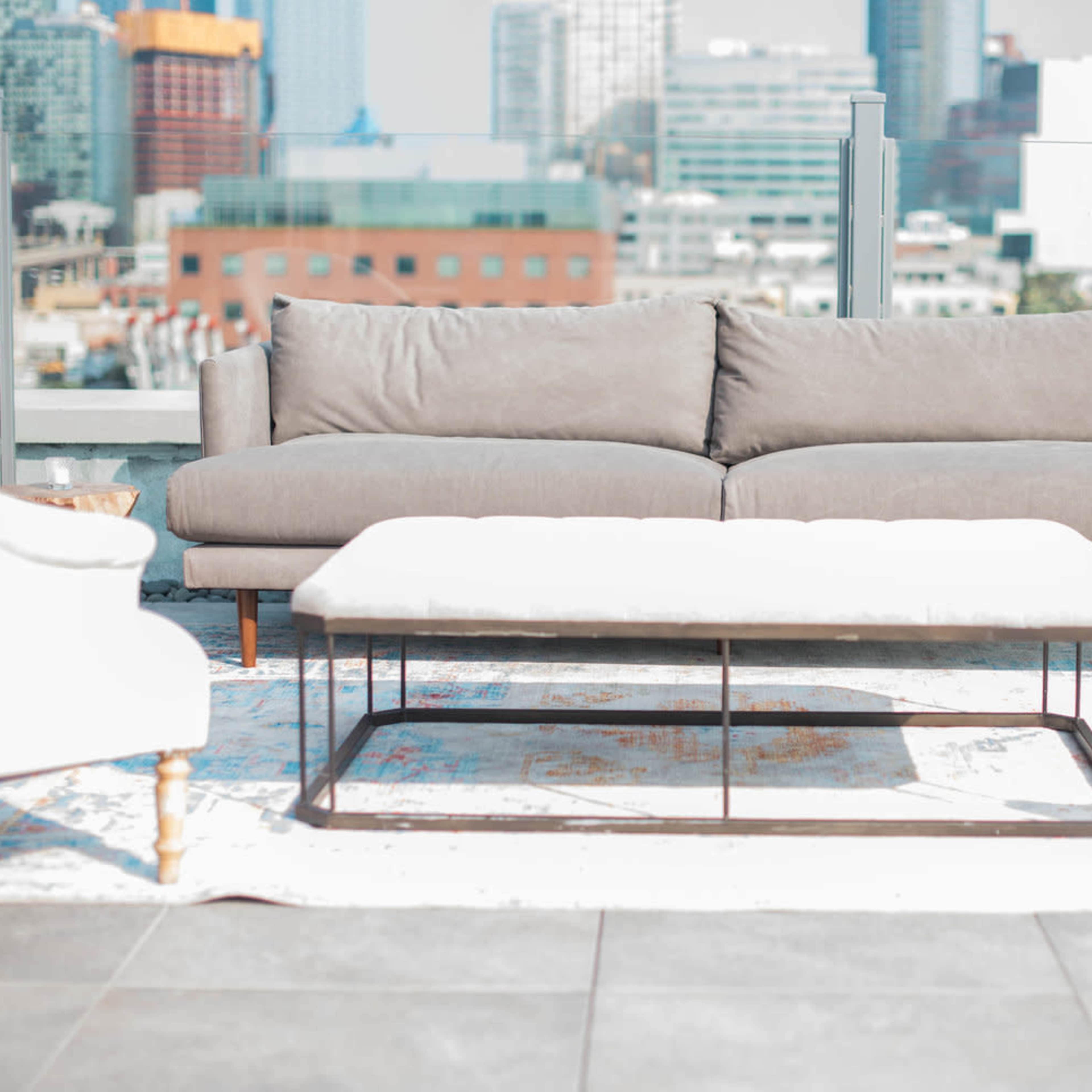 A modern outdoor lounge area features a light gray sofa, a white coffee table, and wooden accent tables on a patterned rug with a city skyline in the background.