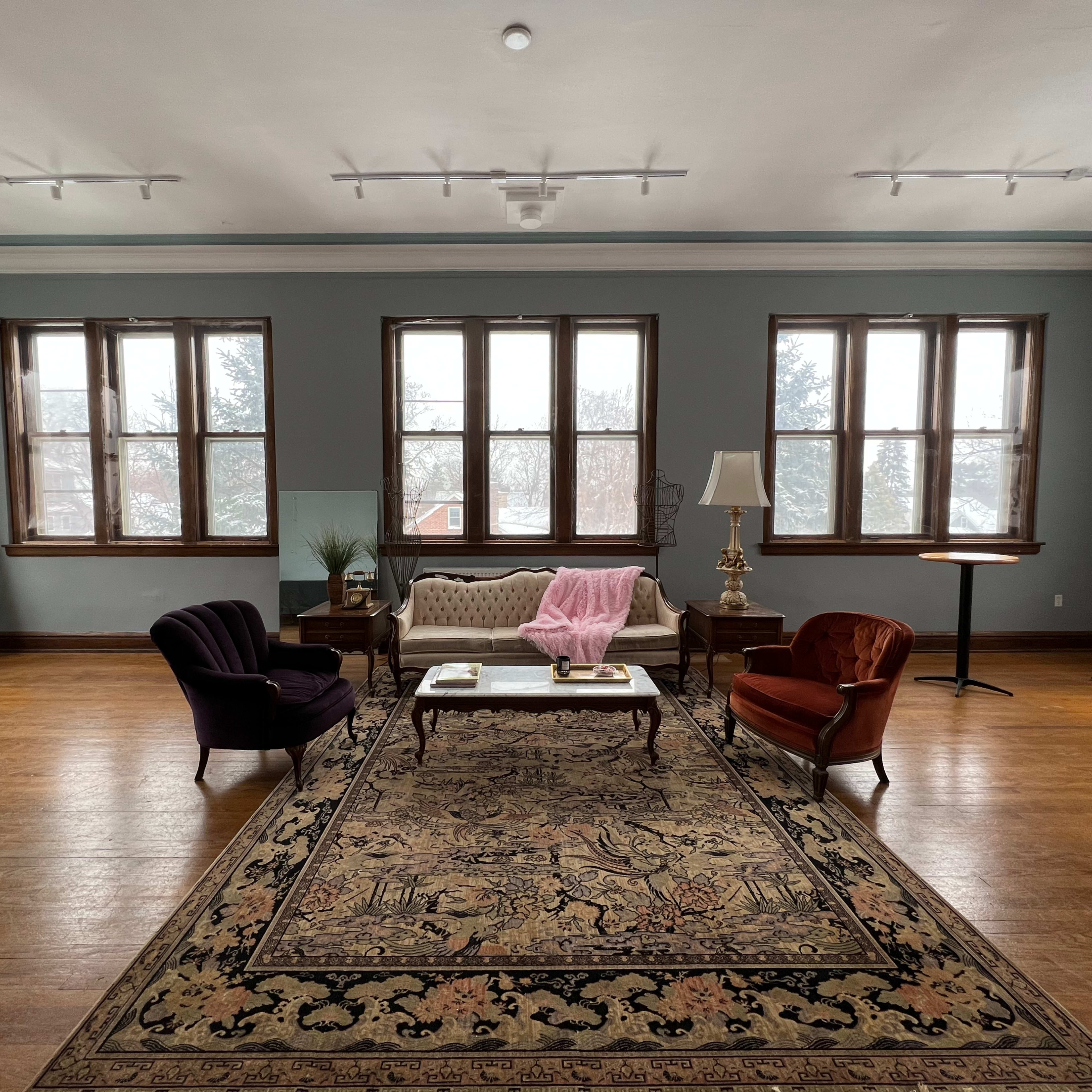 A spacious room features a large area rug with furniture including a couch, two armchairs, a coffee table, and large windows letting in natural light.
