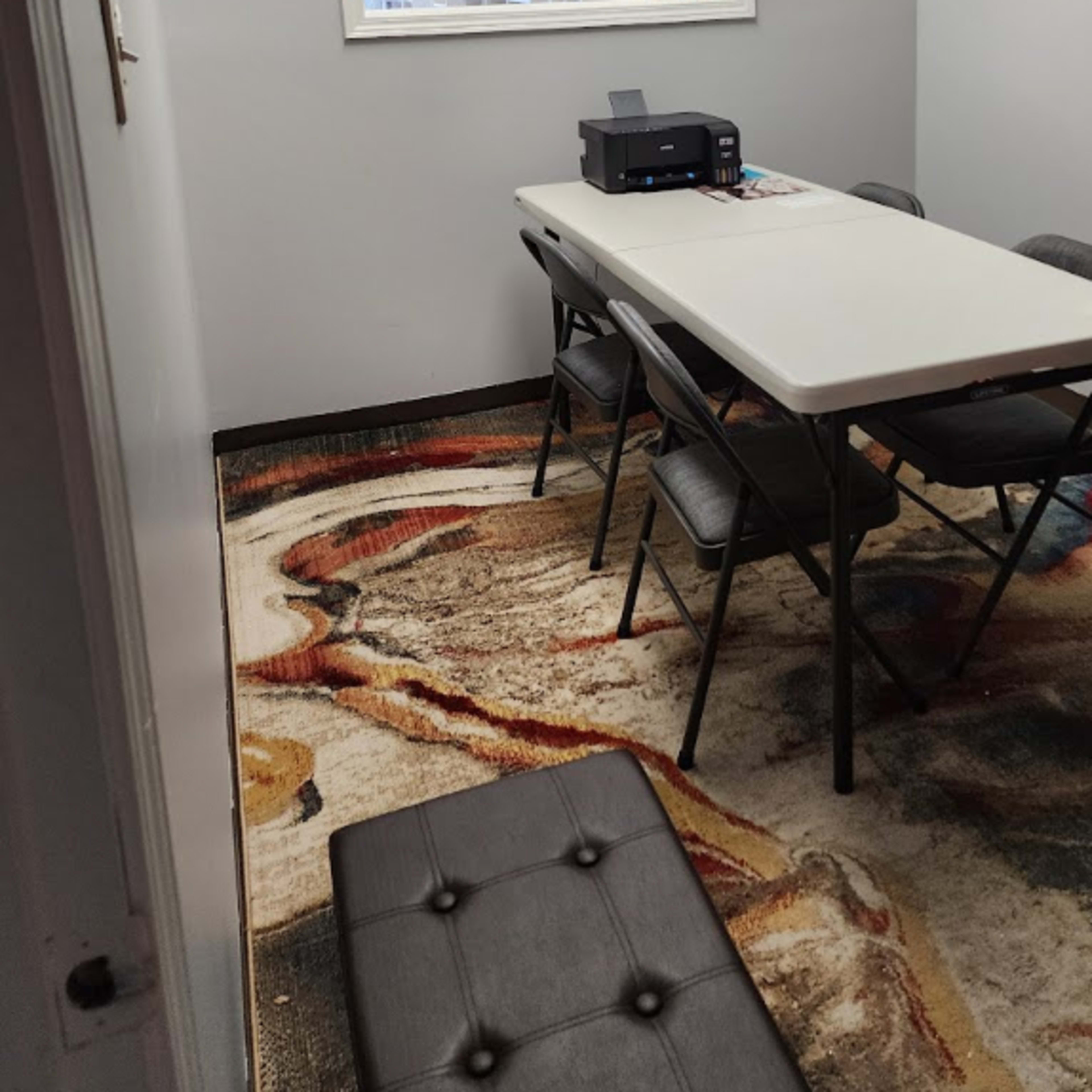 A small meeting room features a table with chairs, a printer, and a large window reflecting a bright space with shelves and plants.