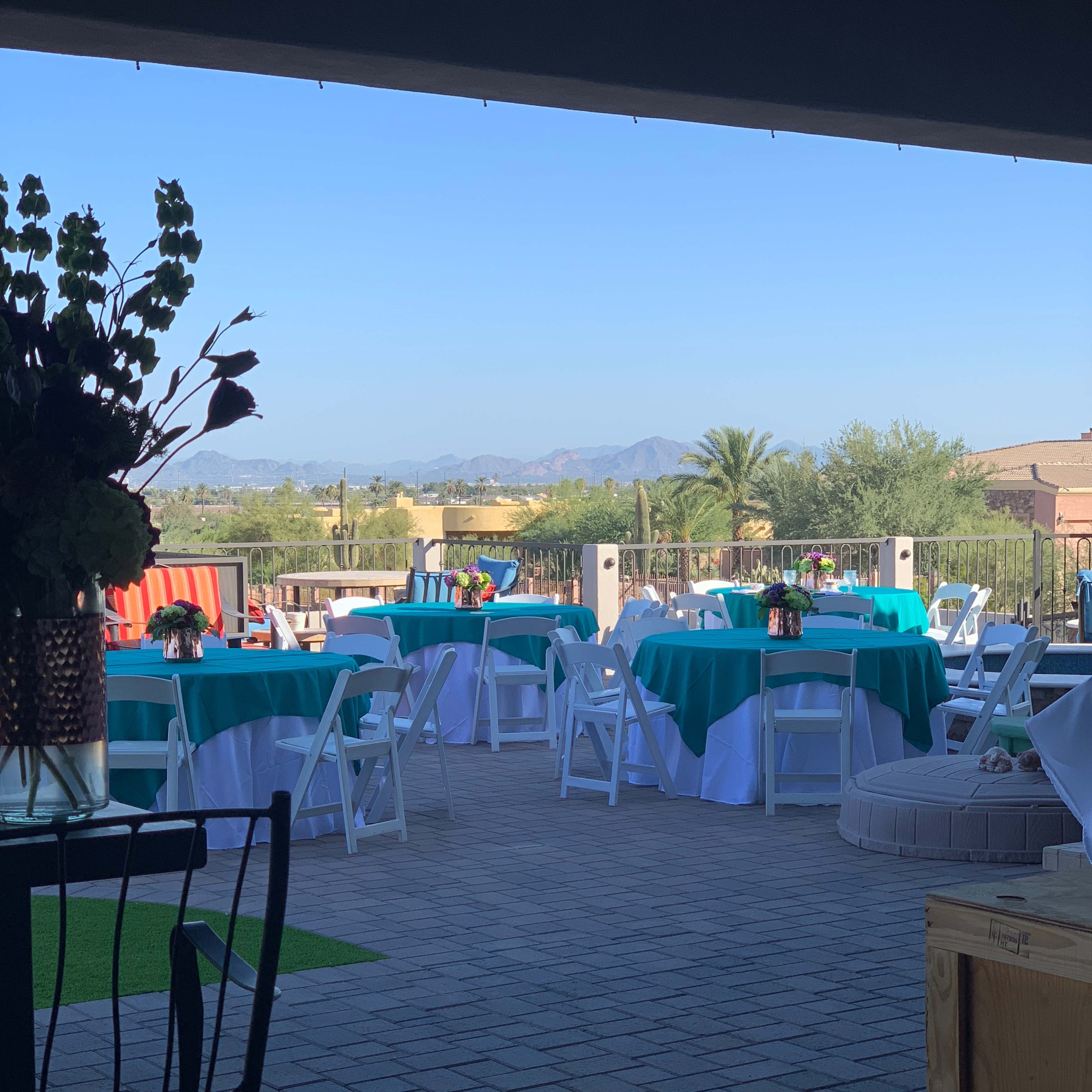 A decorated outdoor event space with tables set up under a patio, surrounded by a scenic view of mountains and greenery.