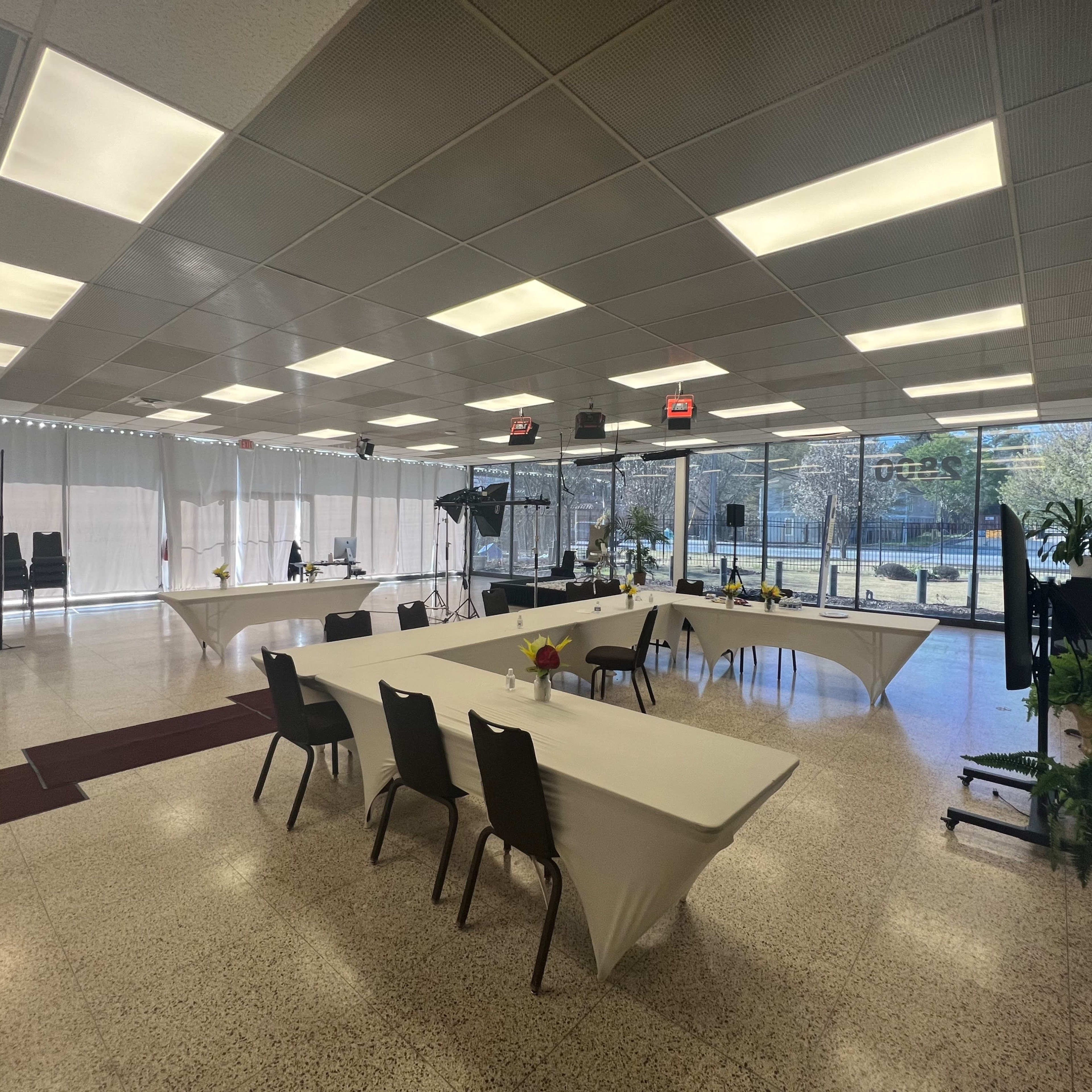 A spacious room is set up with several tables covered in white linens, surrounded by black chairs, and large windows that let in natural light.