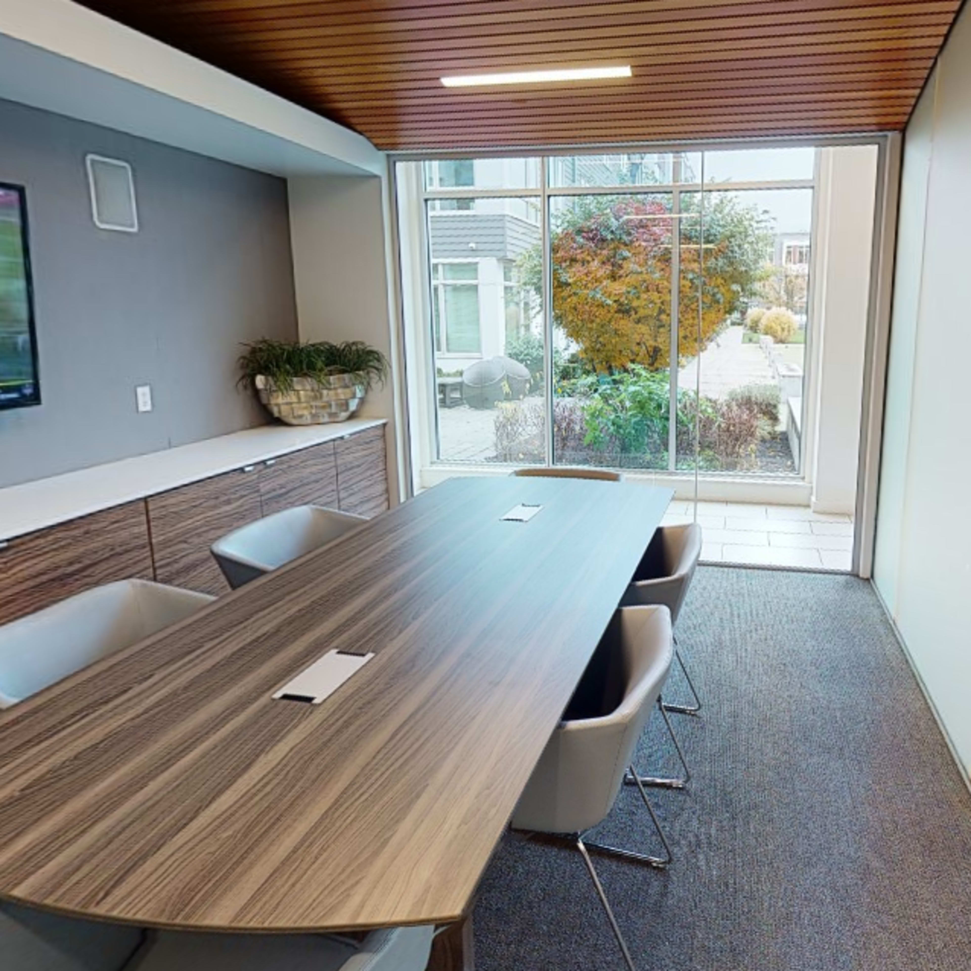 A modern conference room features a large wooden table, six white chairs, and a wall-mounted TV, with a view of a landscaped outdoor area through a large window.