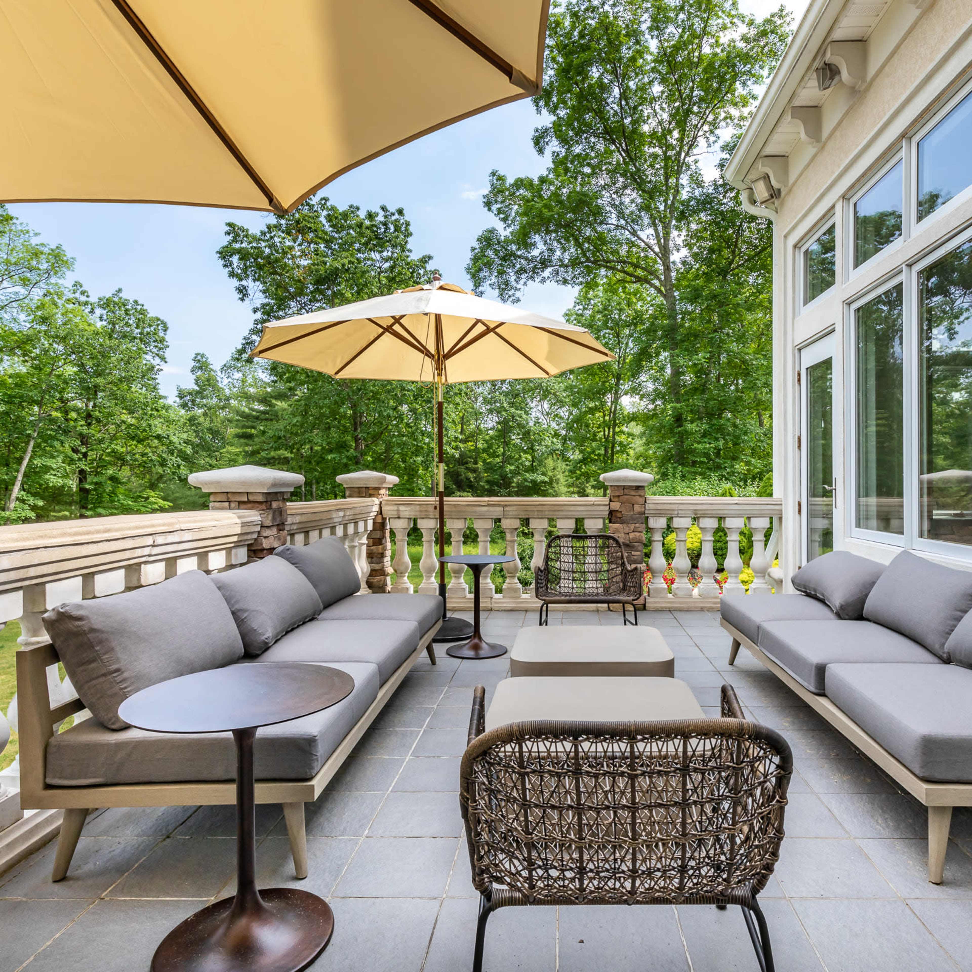 A patio area furnished with grey sofas, a small table, and large umbrellas, surrounded by greenery.