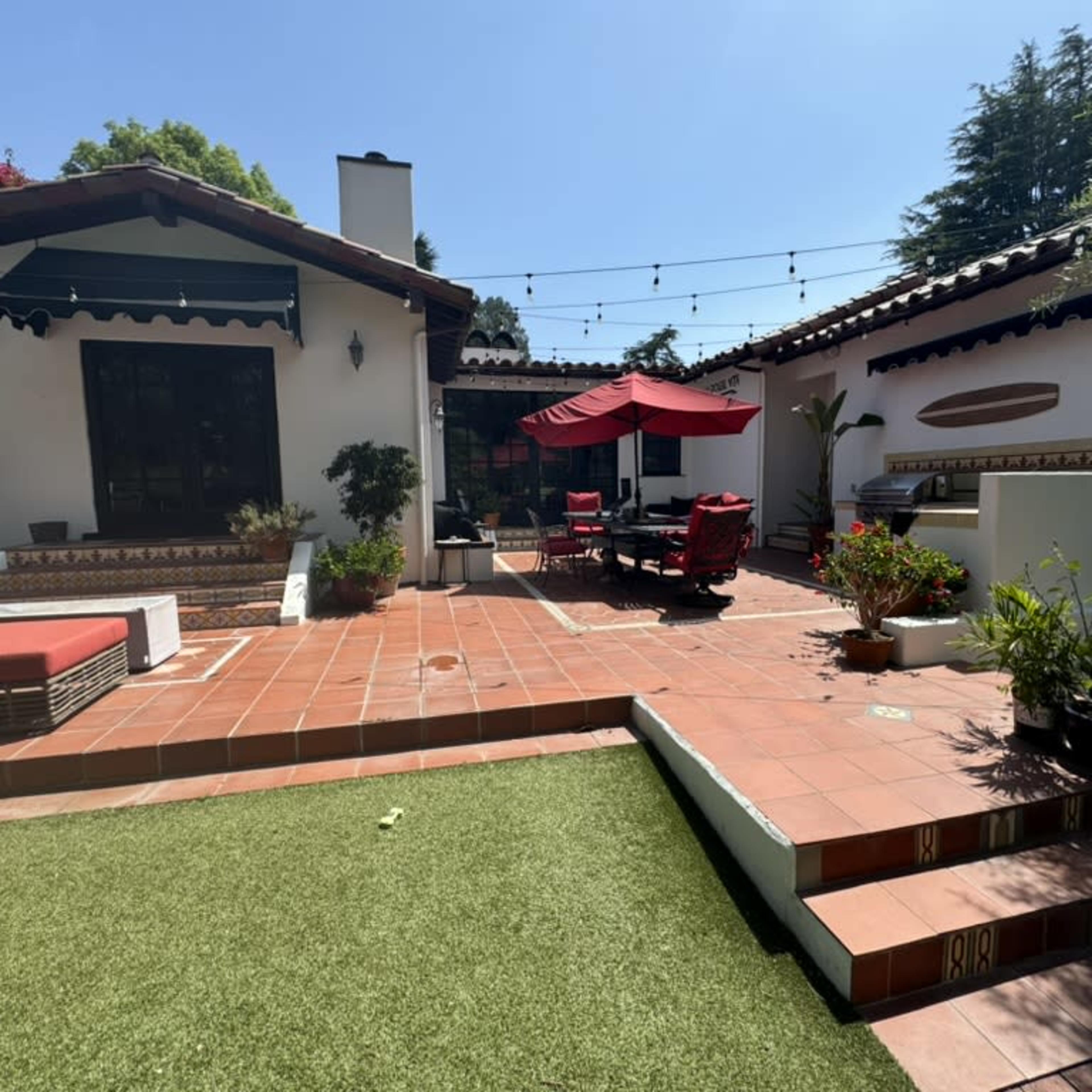 A patio area features terracotta tiles, outdoor seating under a red umbrella, and greenery in pots, with adjacent white-walled buildings.