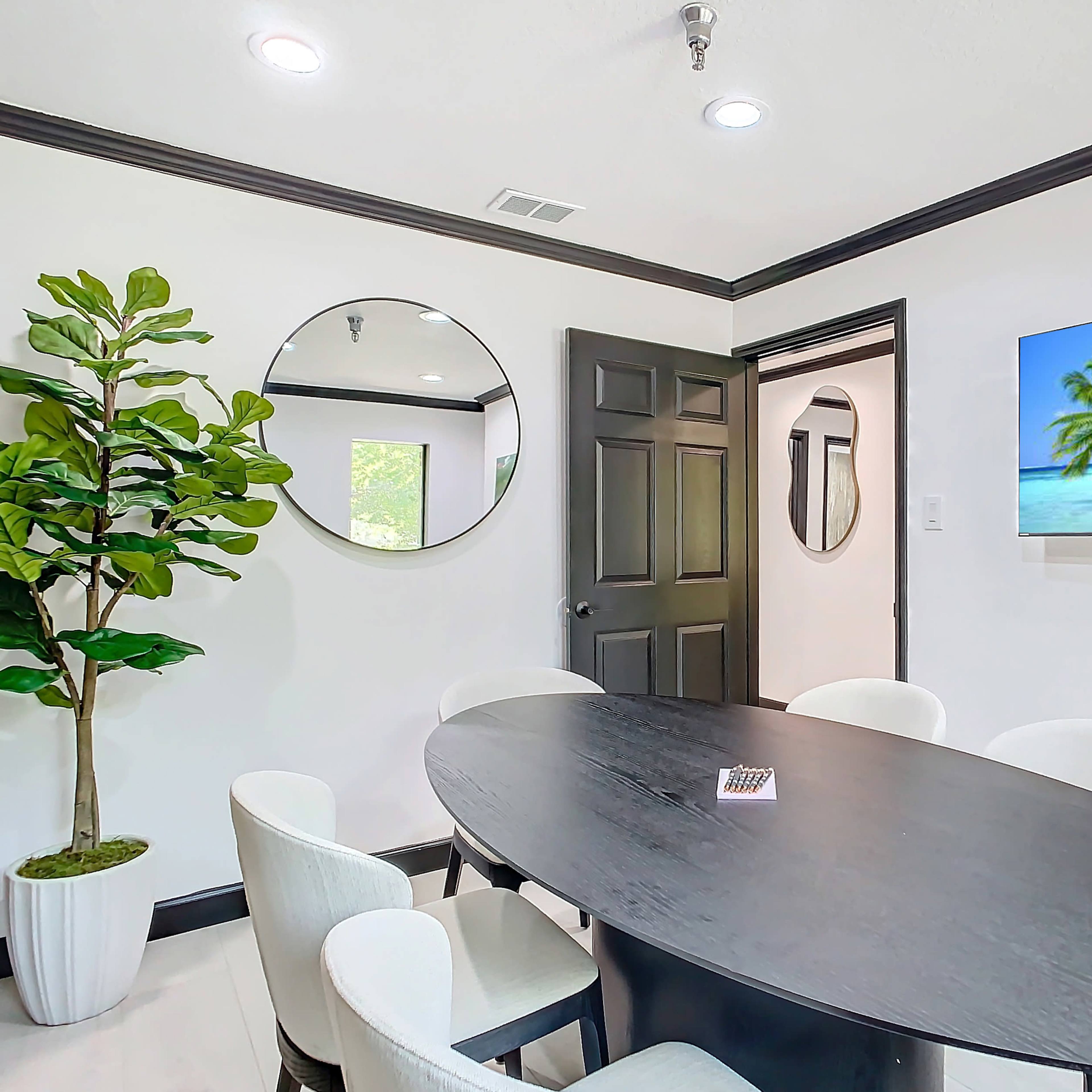 A round black dining table surrounded by white chairs is positioned in a bright room featuring a large mirror and a beach-themed TV display.