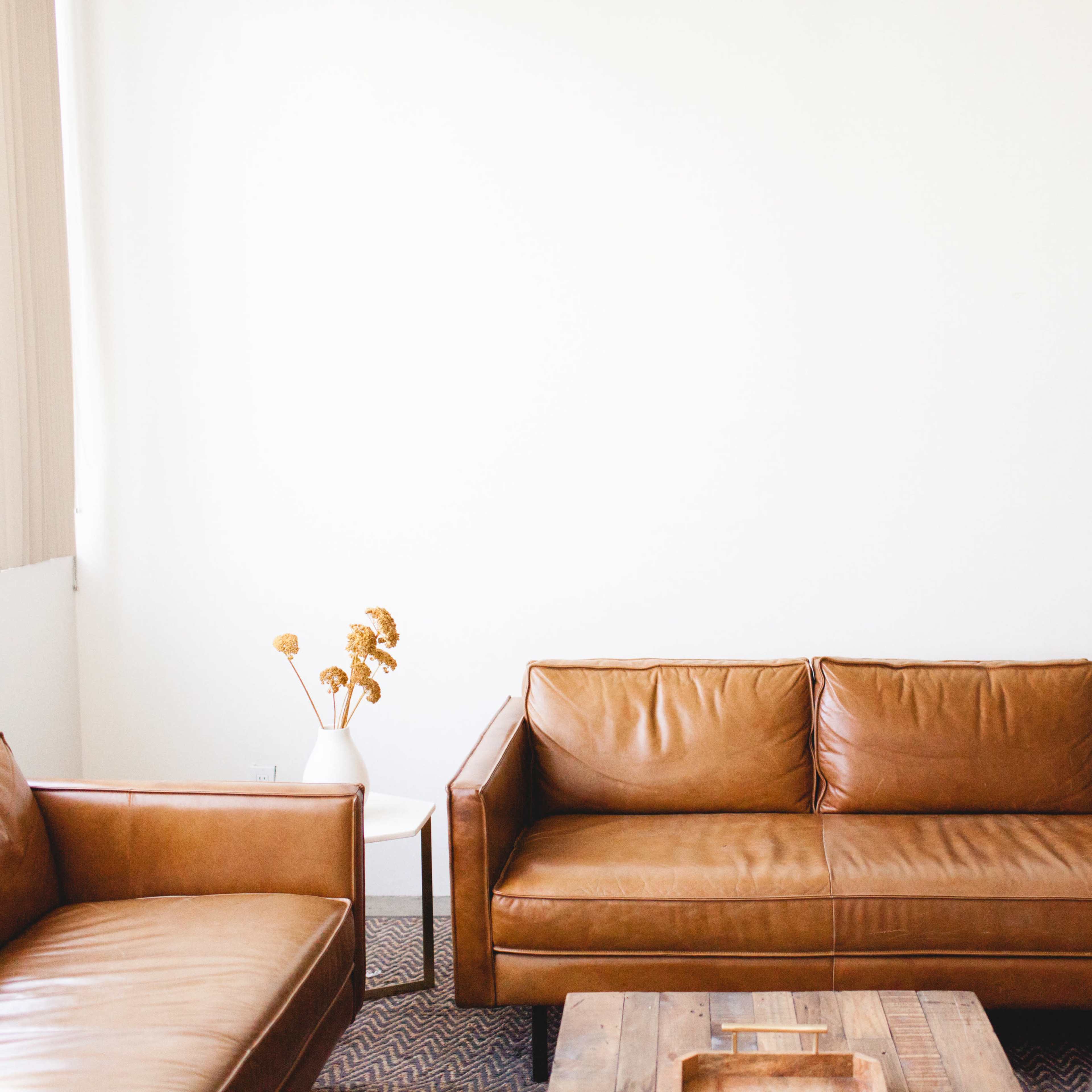 A light-filled room features two brown leather sofas and a wooden coffee table, along with a vase containing dried flowers.