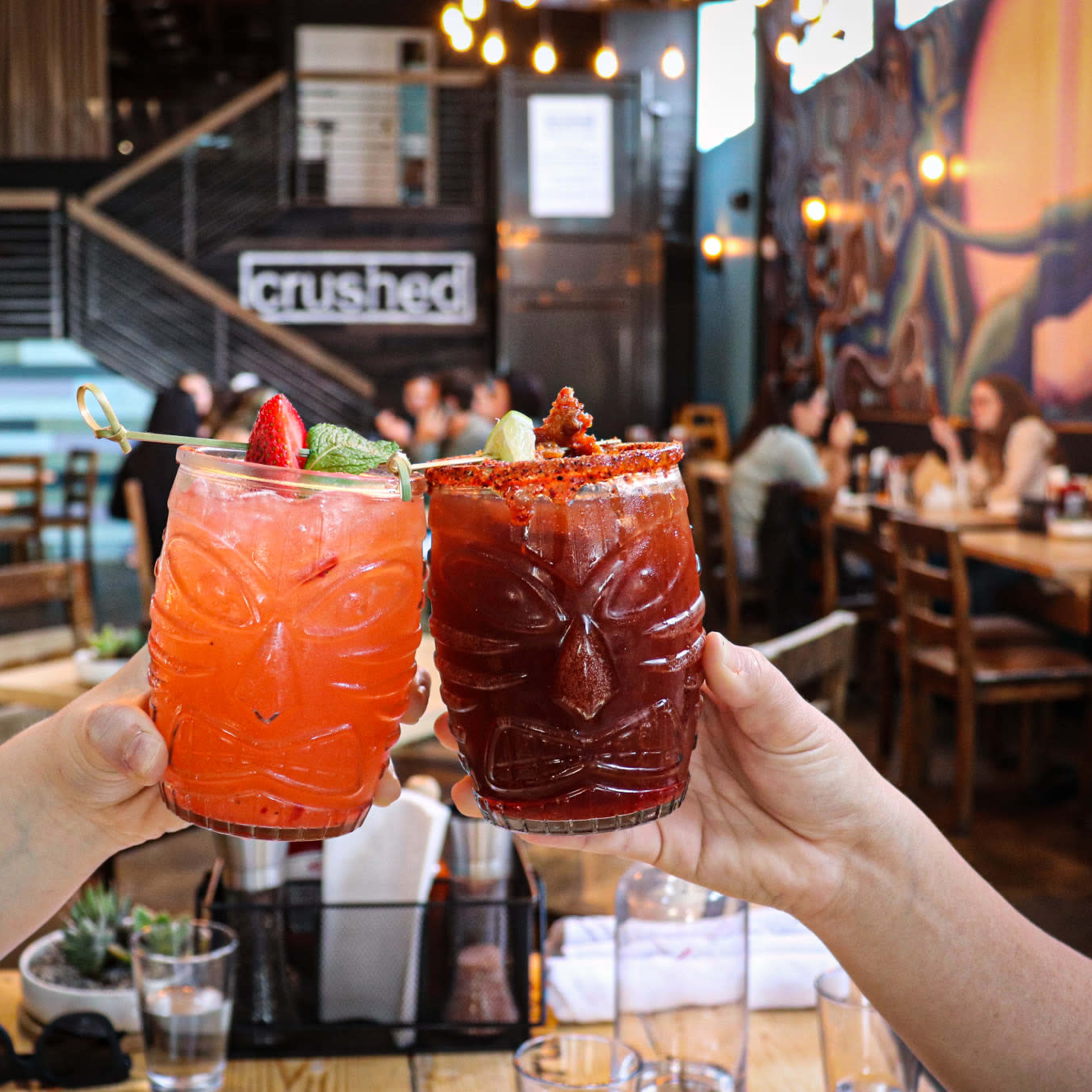 Two colorful cocktails in decorative glasses are being clinked together in a lively restaurant setting.
