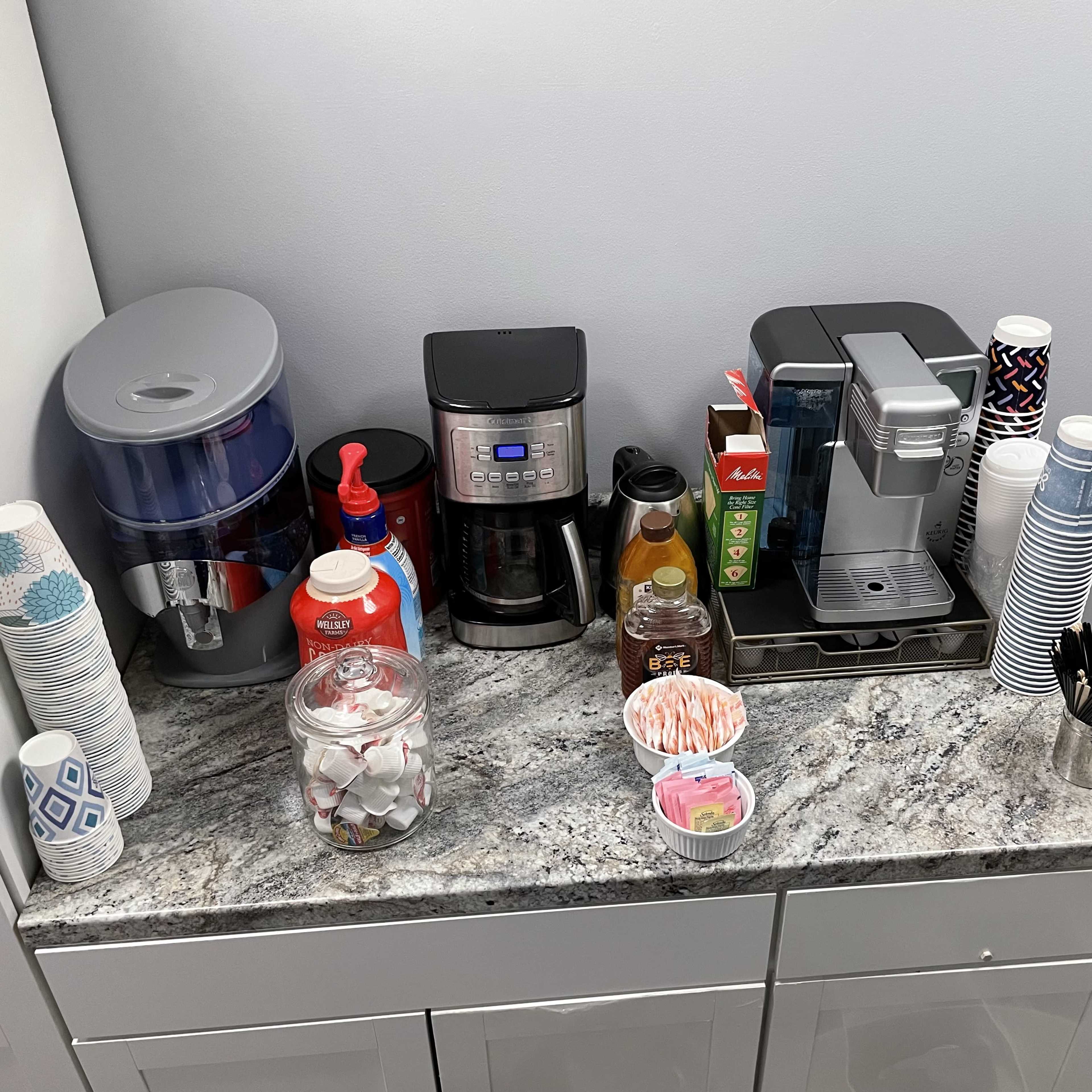 A countertop with a water dispenser, a coffee maker, an espresso machine, various cups, a jar of sugar packets, and condiments neatly arranged.