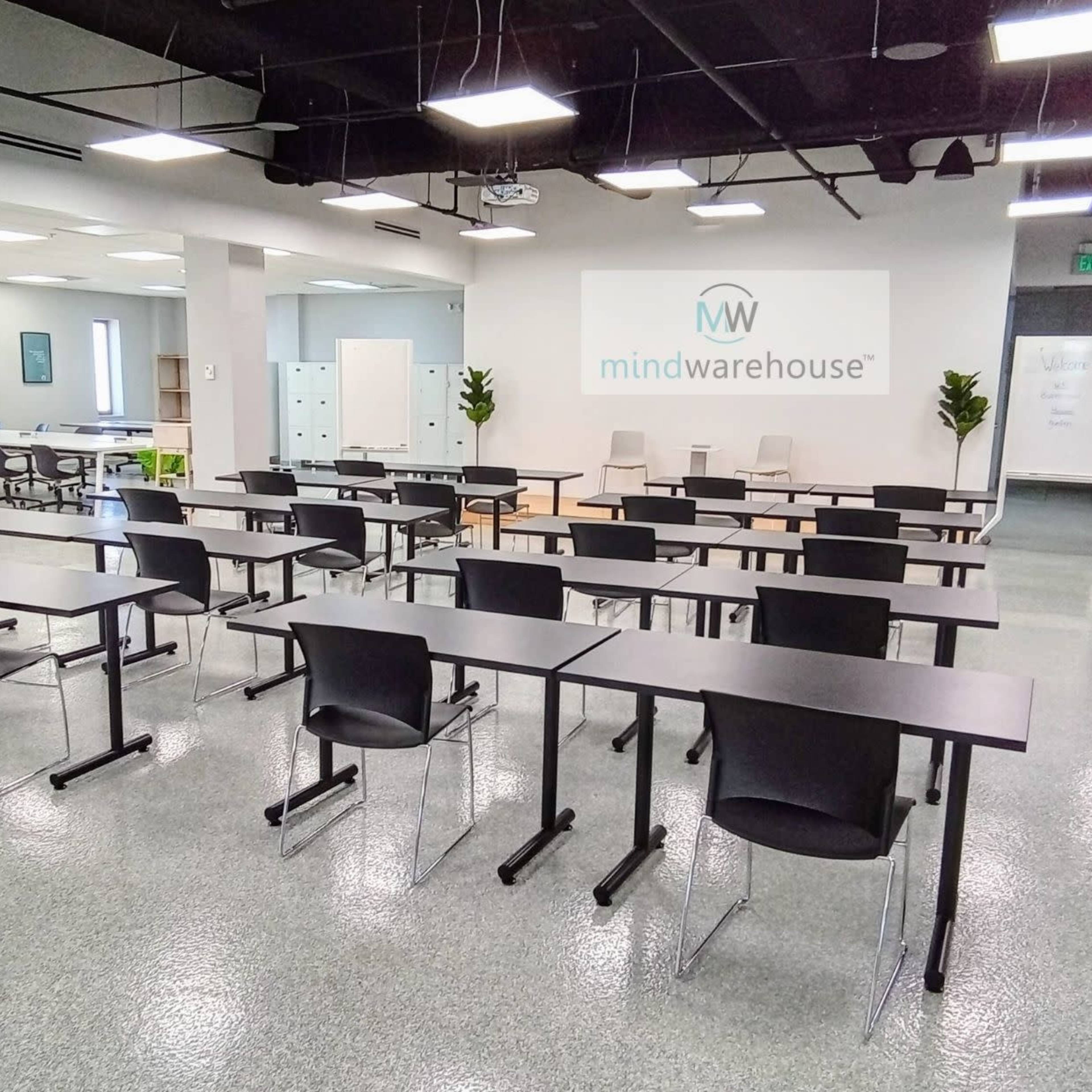 A modern classroom or meeting space featuring rows of black tables and chairs, with a minimalistic design and a logo on the back wall.