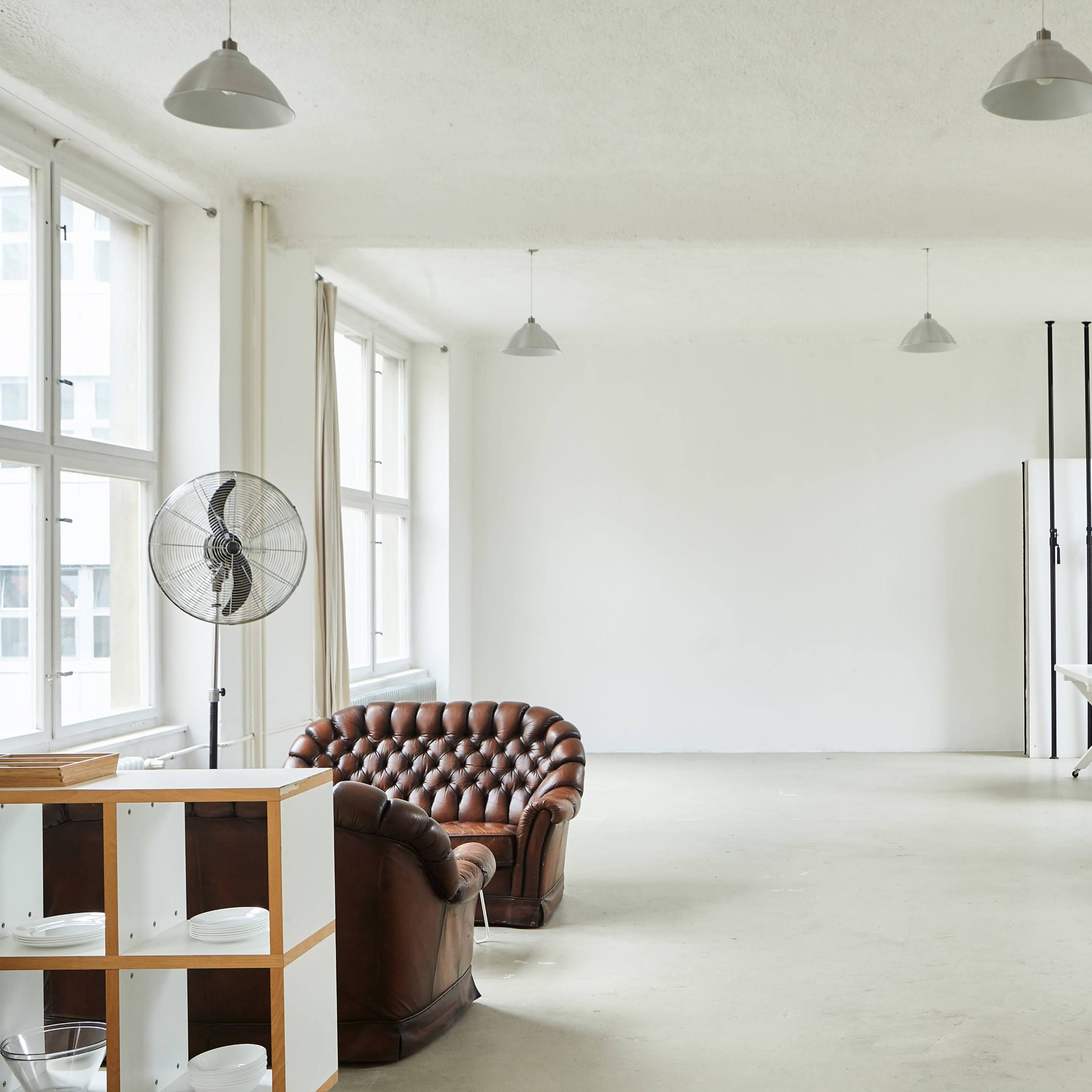 A spacious, minimalistic room features a brown leather couch, a wooden table, a fan, and simple furnishings against white walls and large windows.