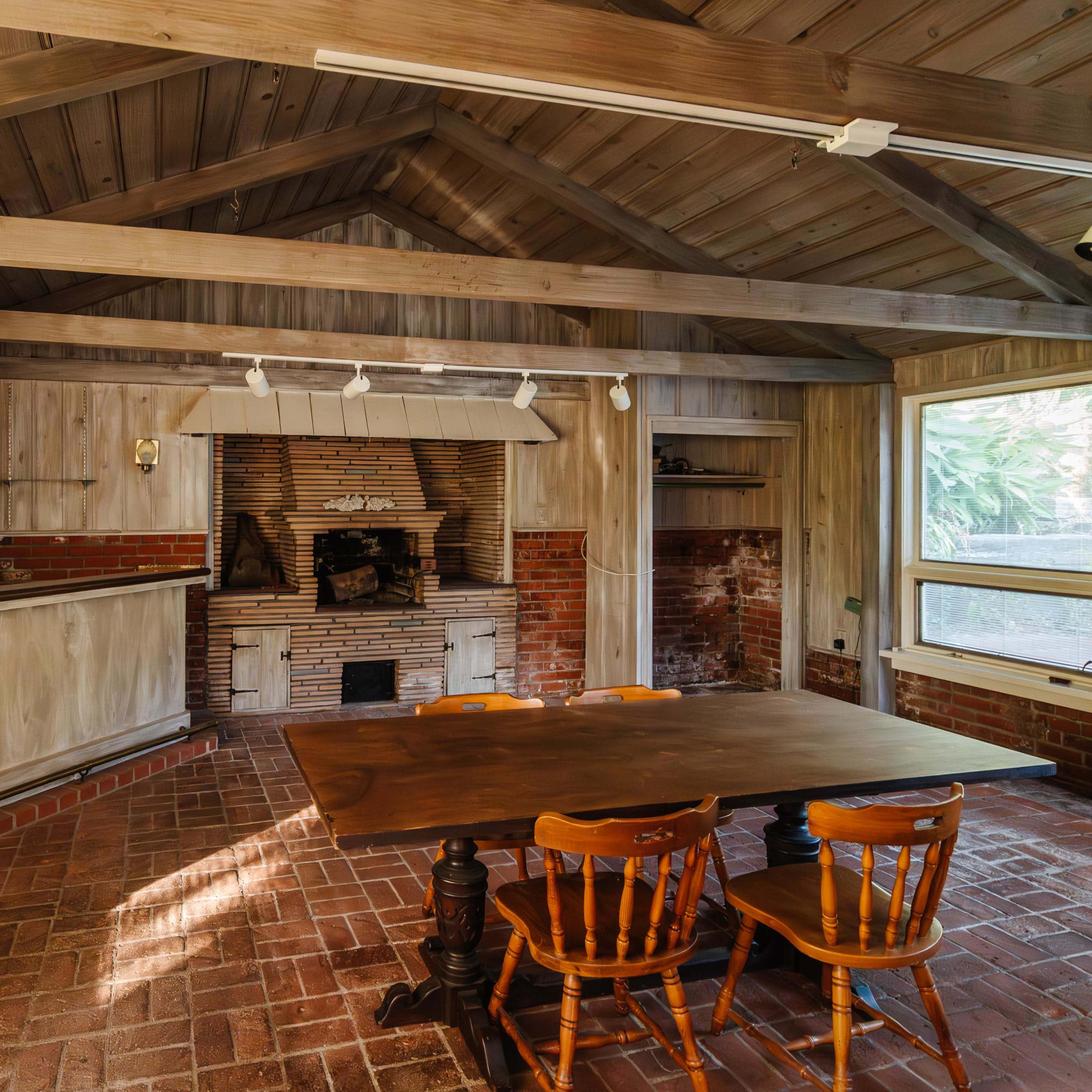 A rustic dining area with a wooden table and chairs, adjacent to a brick fireplace and surrounded by large windows.