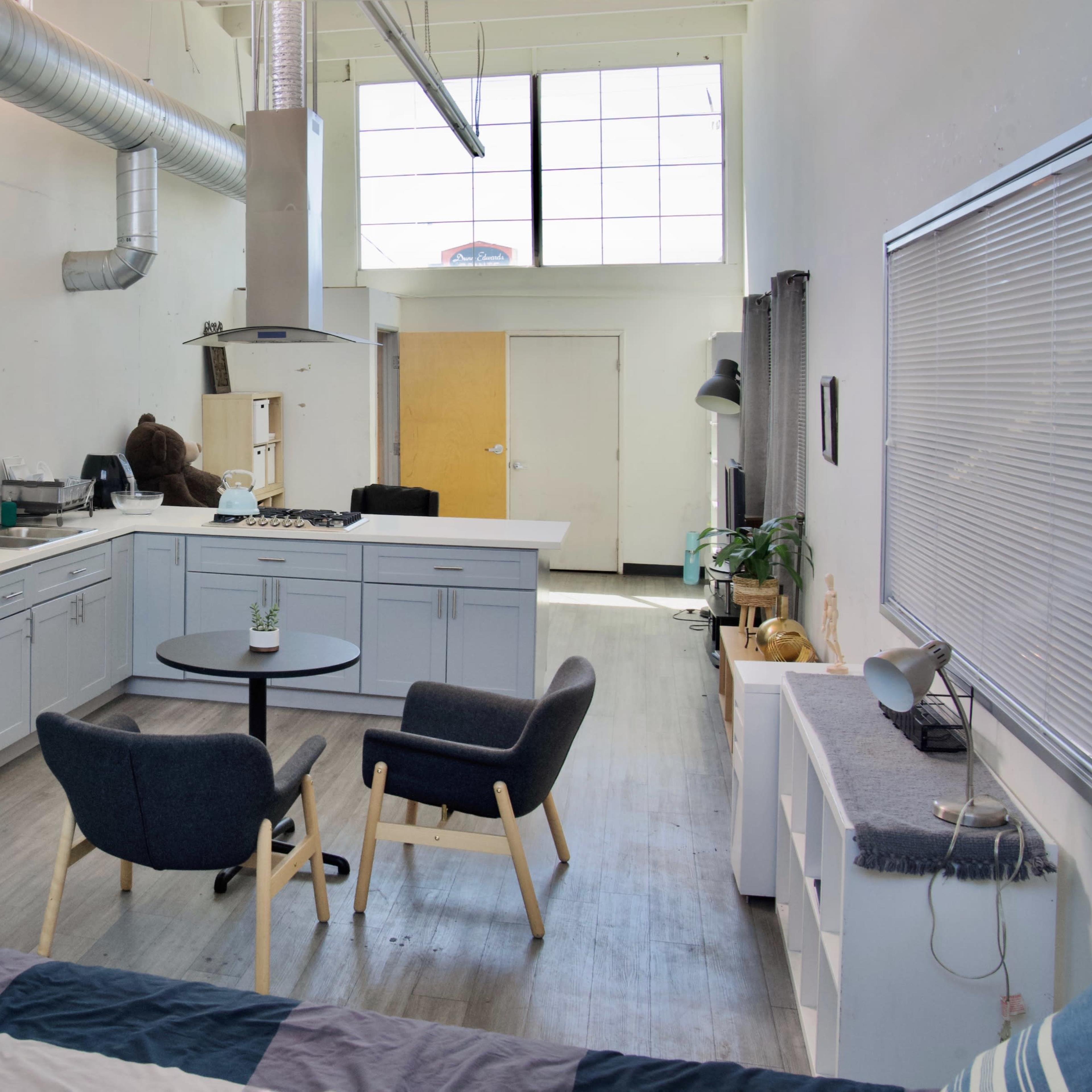 A modern kitchen and living area with a refrigerator, a table with chairs, and large windows allowing natural light.