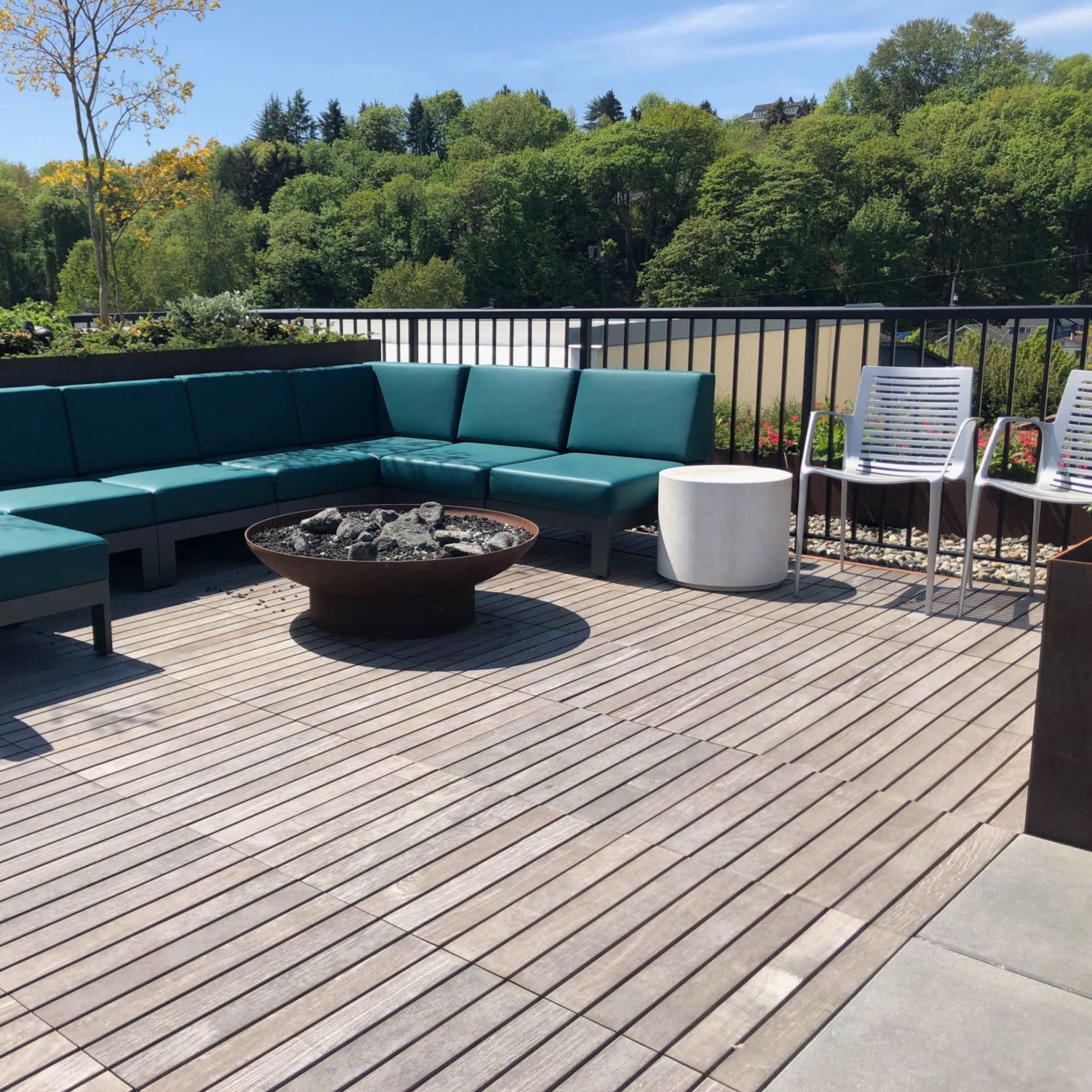 A modern outdoor seating area features a turquoise sectional sofa surrounding a round fire pit on a wooden deck, with a white table and additional chairs nearby.
