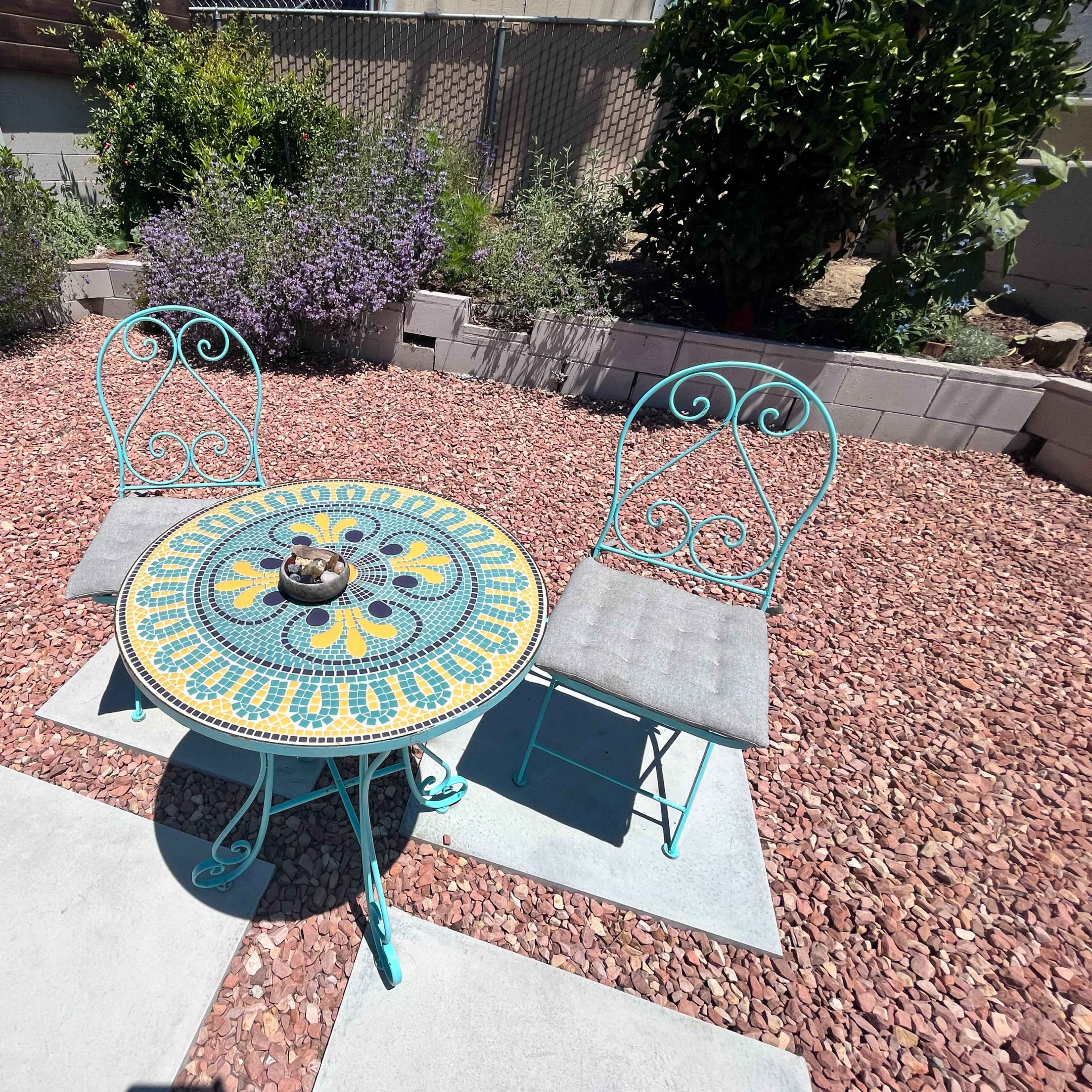 A round, intricately designed table with two blue metal chairs is set up on a gravel patio surrounded by lush plants.