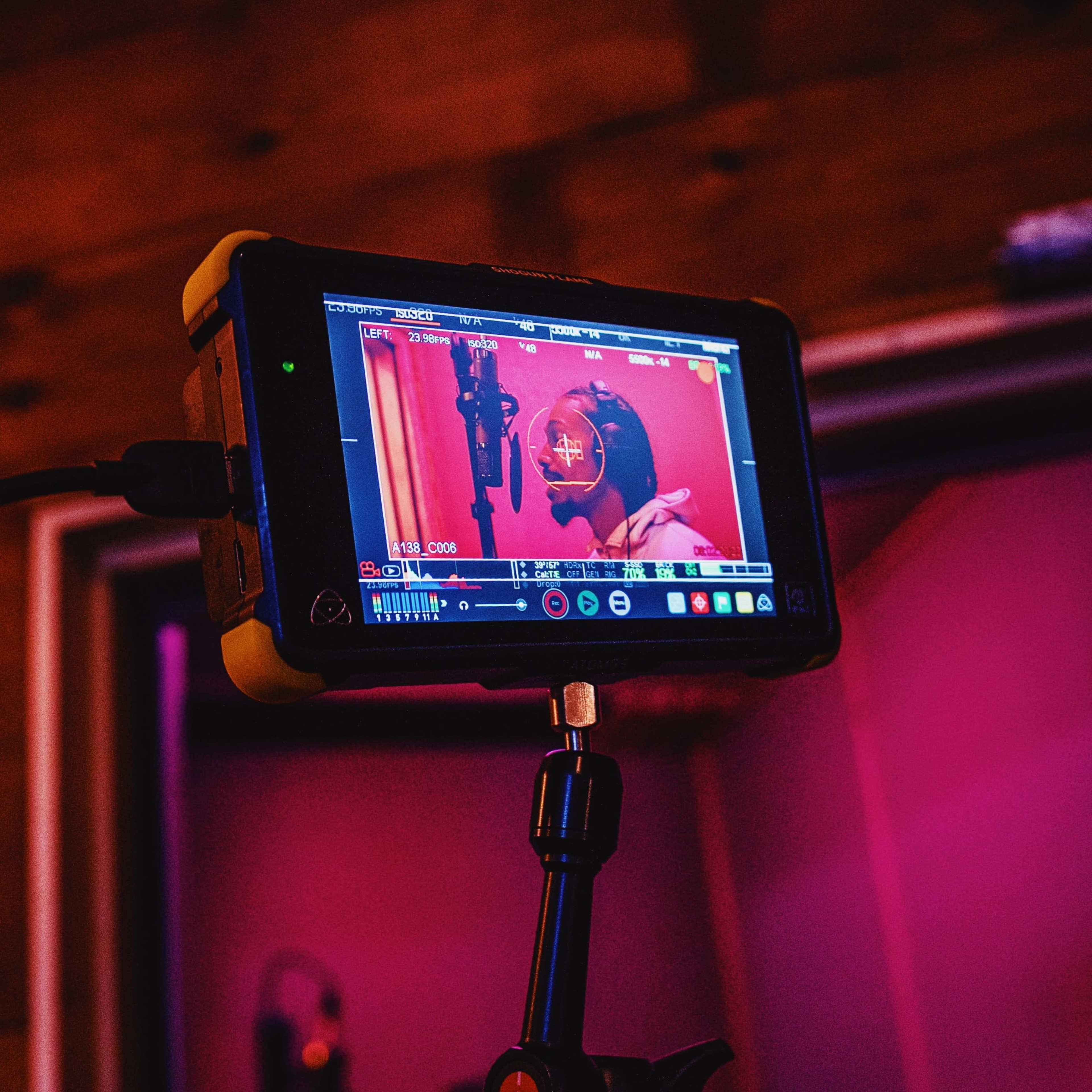 A monitor displaying a recording session in a sound studio is positioned on a stand, showing a microphone and a performer in front of a pink-lit background.