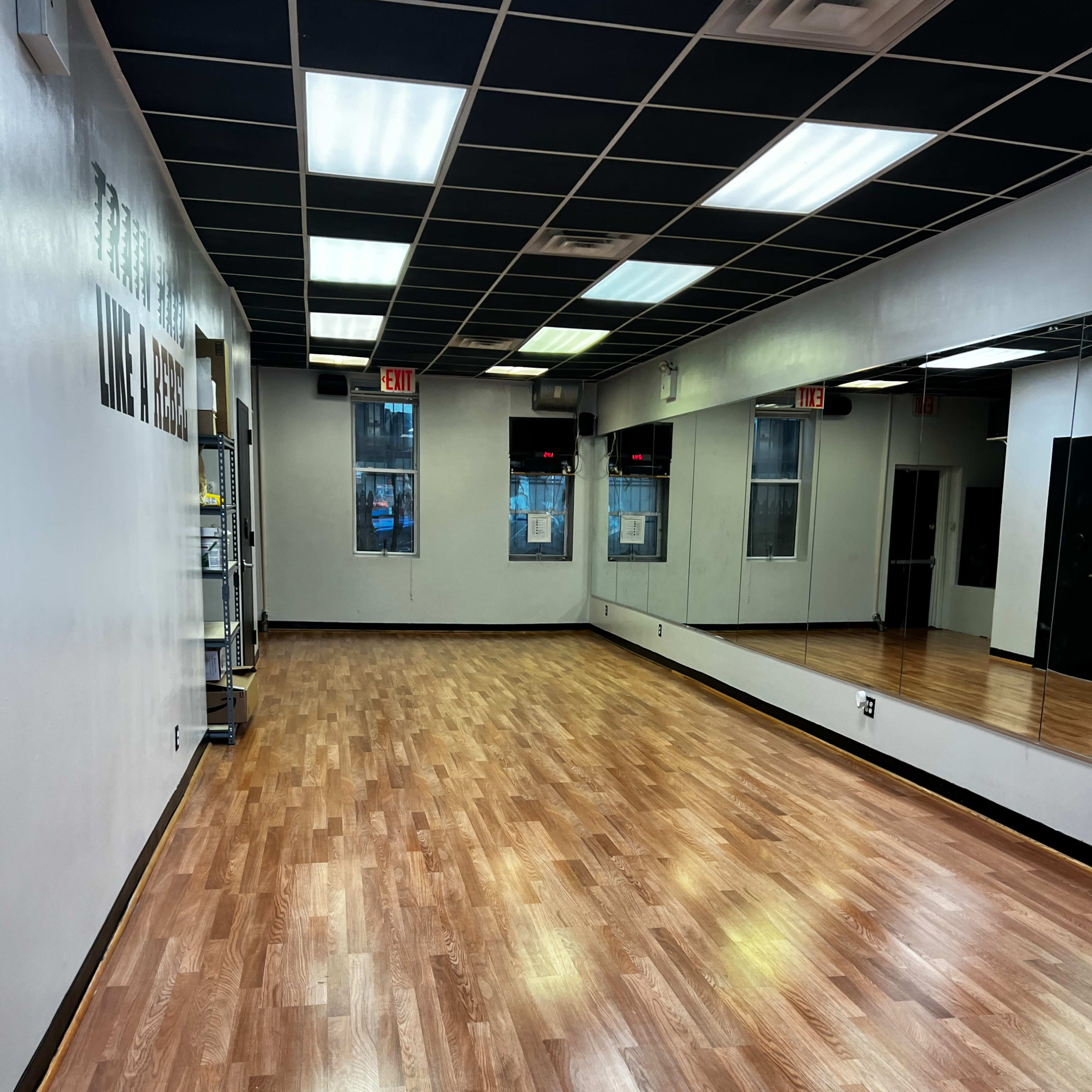 An empty studio with hardwood flooring and mirrored walls, illuminated by overhead lights.