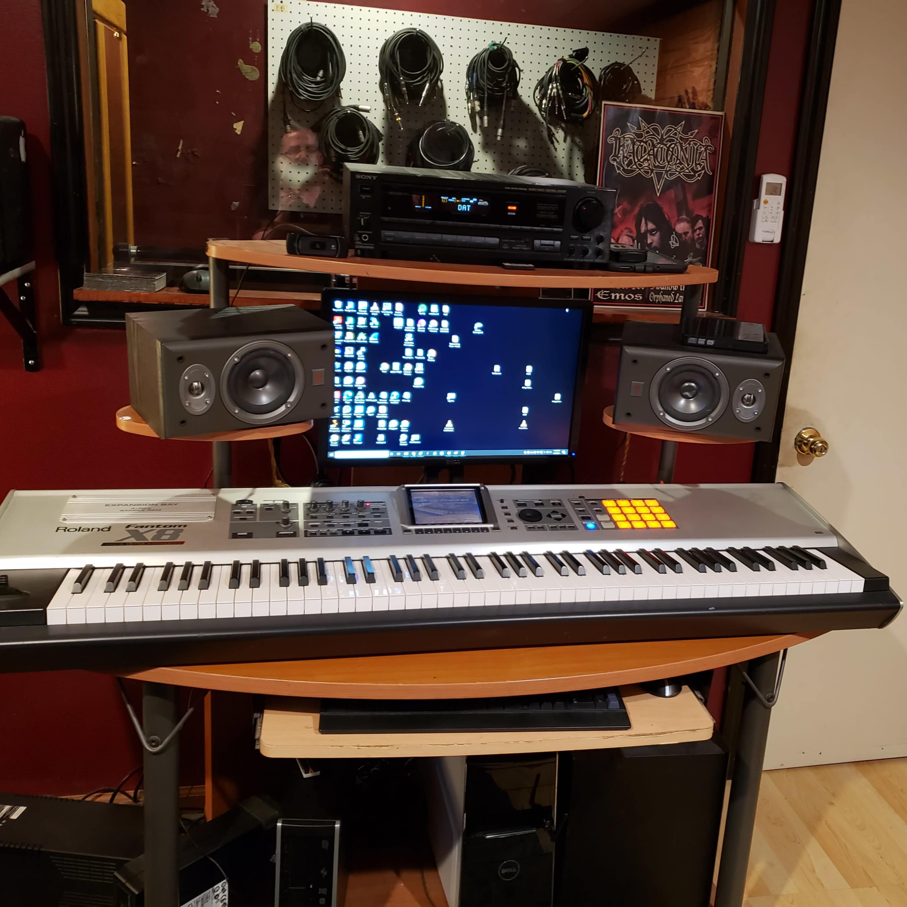 A music production setup featuring a keyboard, computer monitor, and audio speakers arranged on a desk against a red wall.