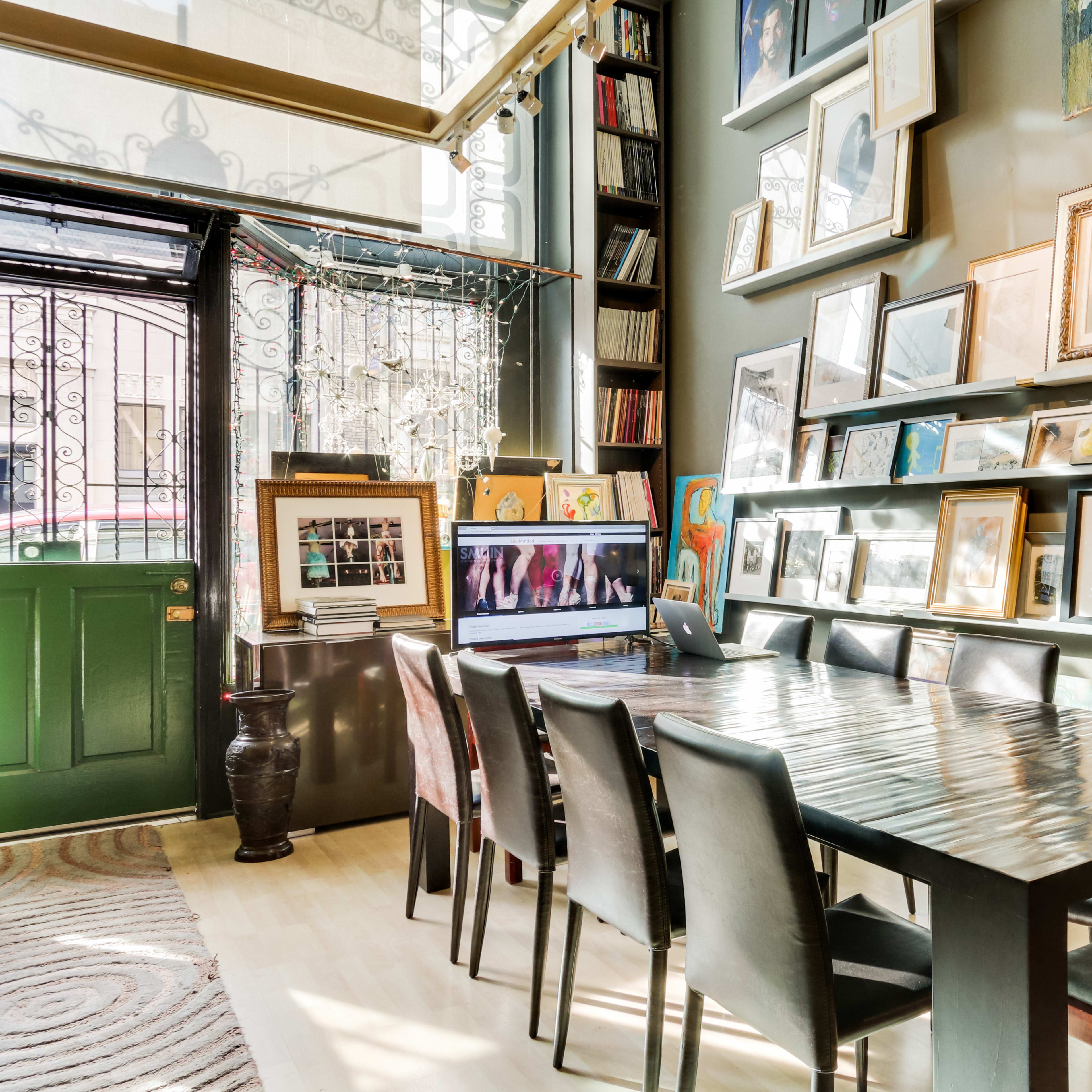 A long wooden table with several black chairs is set in a room filled with framed photographs and artwork on the walls, with large windows letting in natural light.