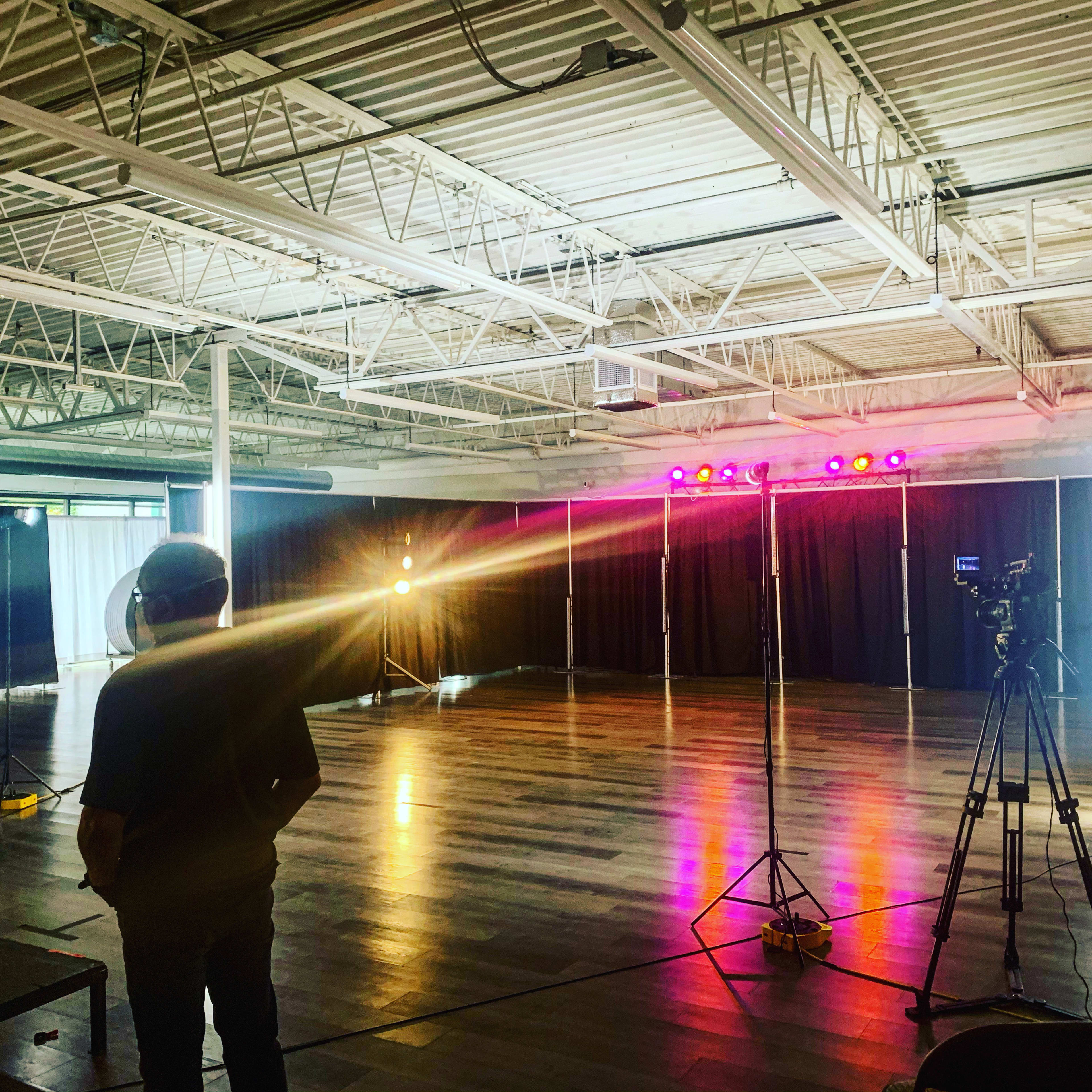 A person stands in an empty studio with colored lights set up and a camera facing a backdrop.
