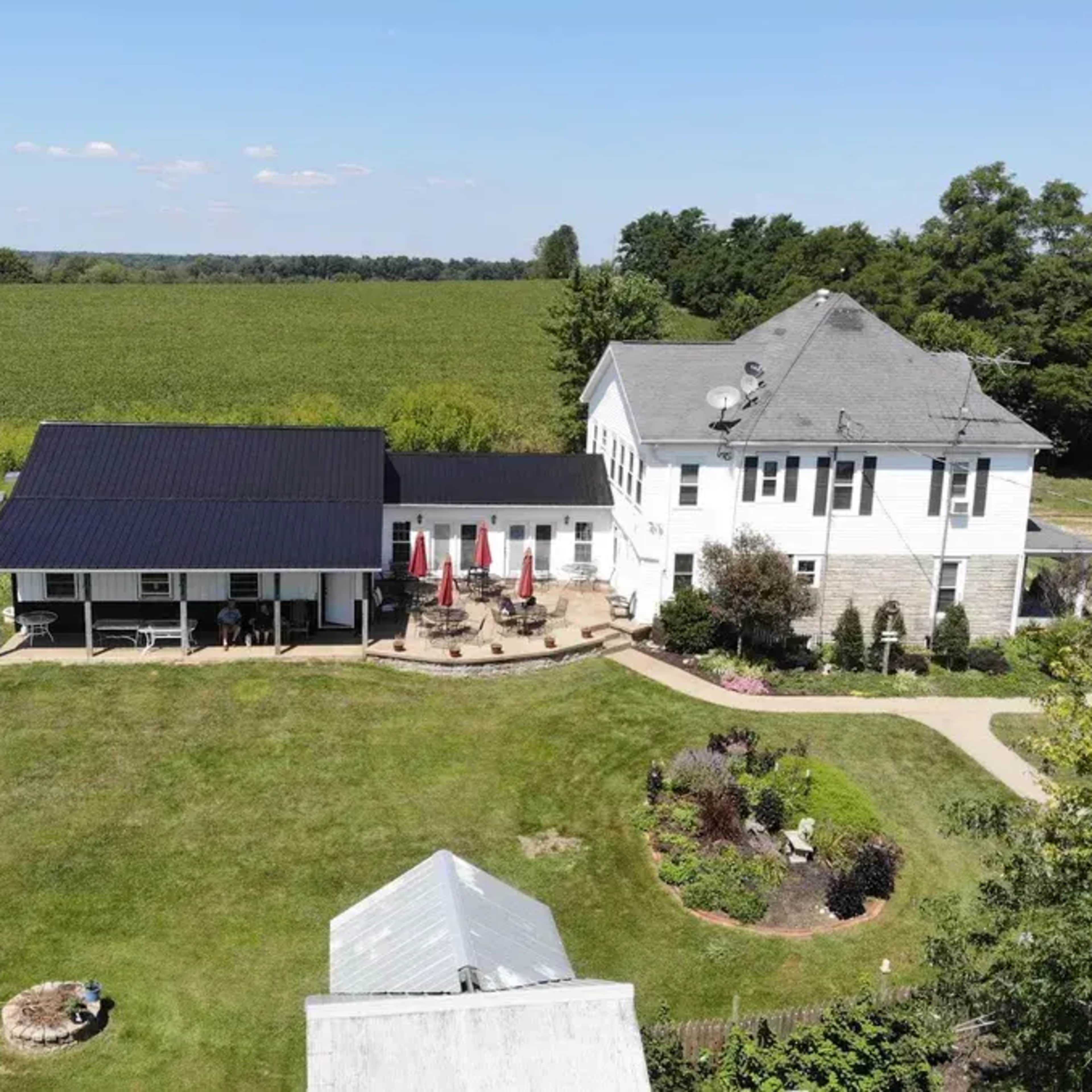 A large two-story house with a black roof and an adjoining single-story structure, surrounded by a well-maintained lawn and a field in the background.