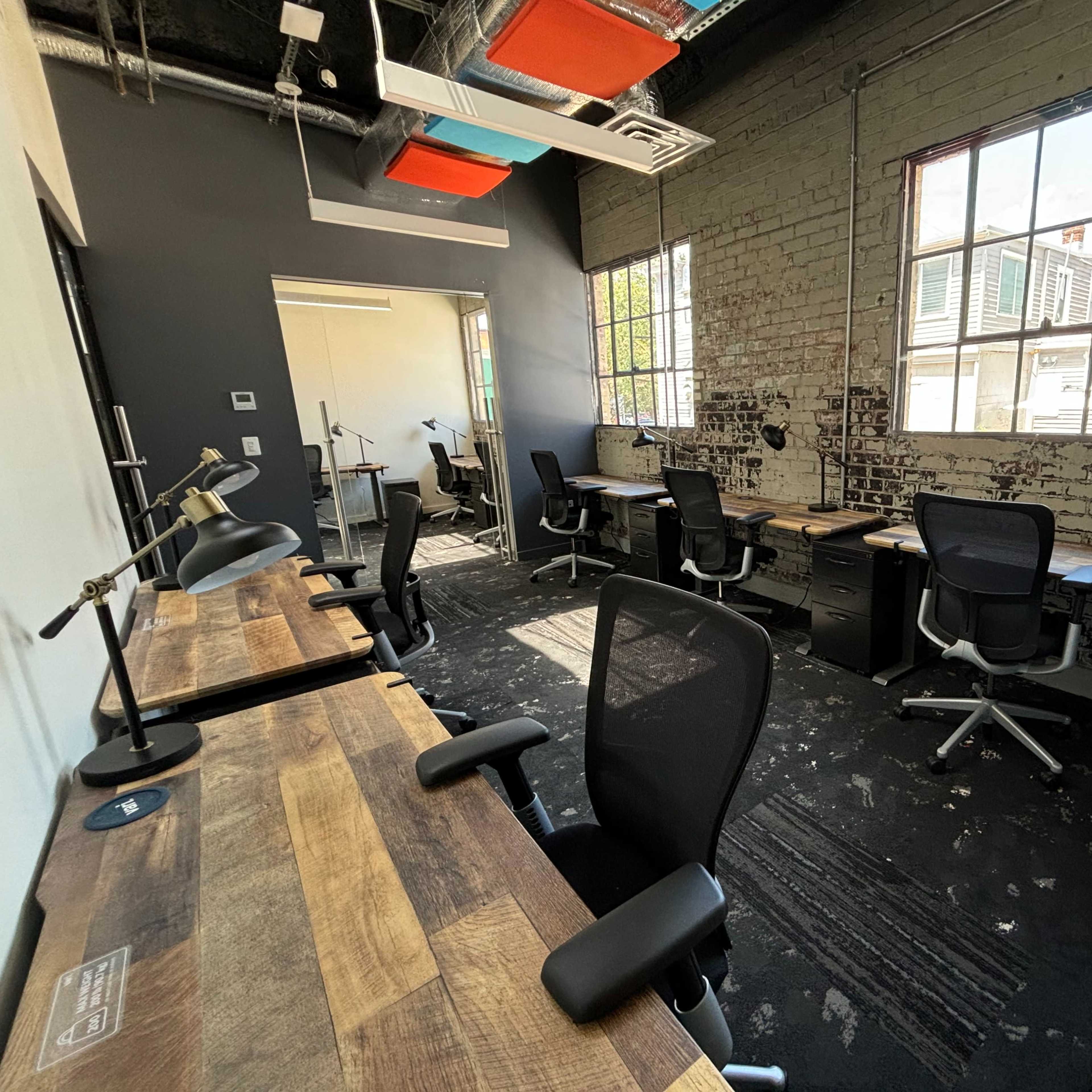 A spacious office with several desks and chairs arranged near large windows, along with exposed brick walls and industrial lighting.