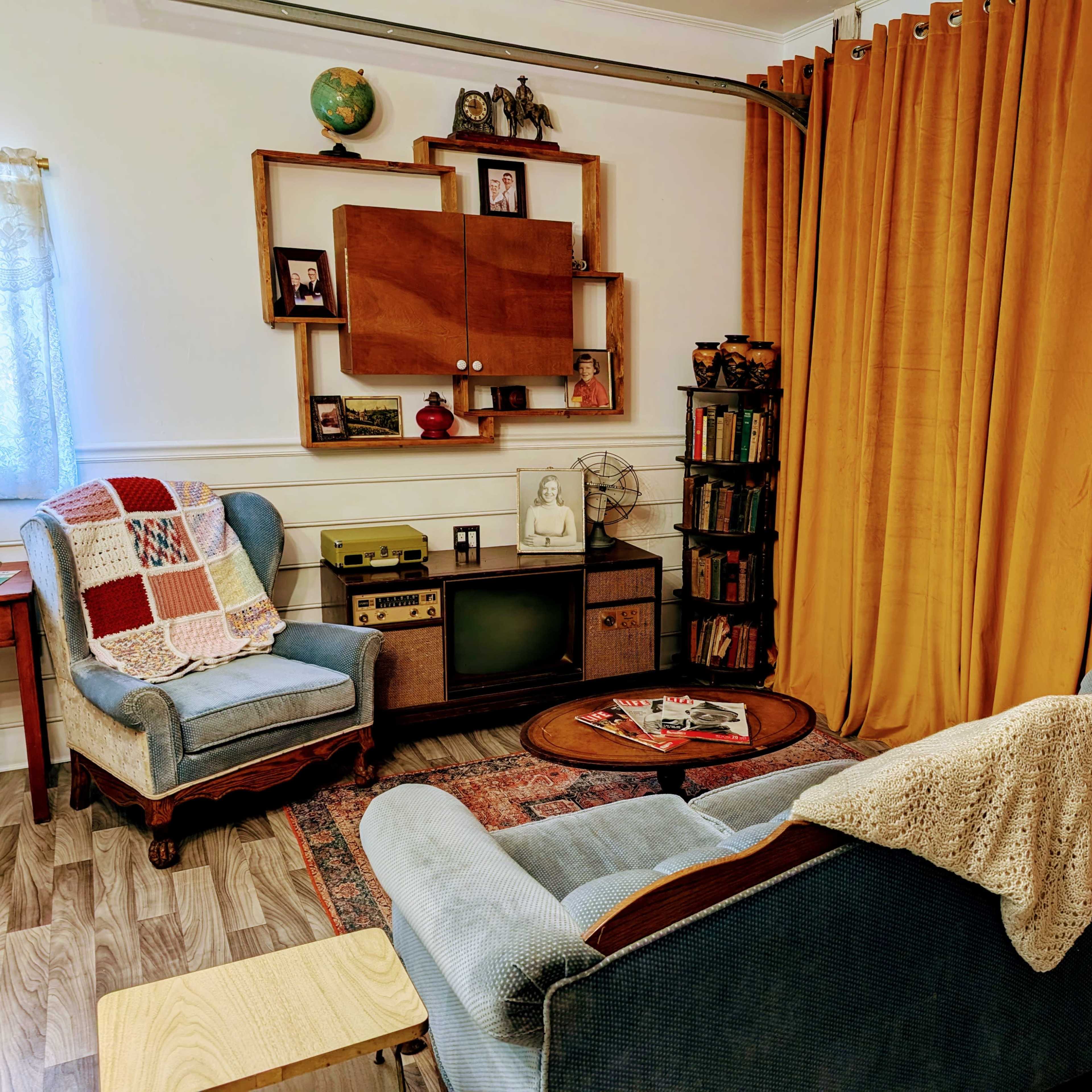 A cozy living room with two upholstered chairs, a wooden coffee table, a vintage television, shelves filled with books, and yellow curtains.