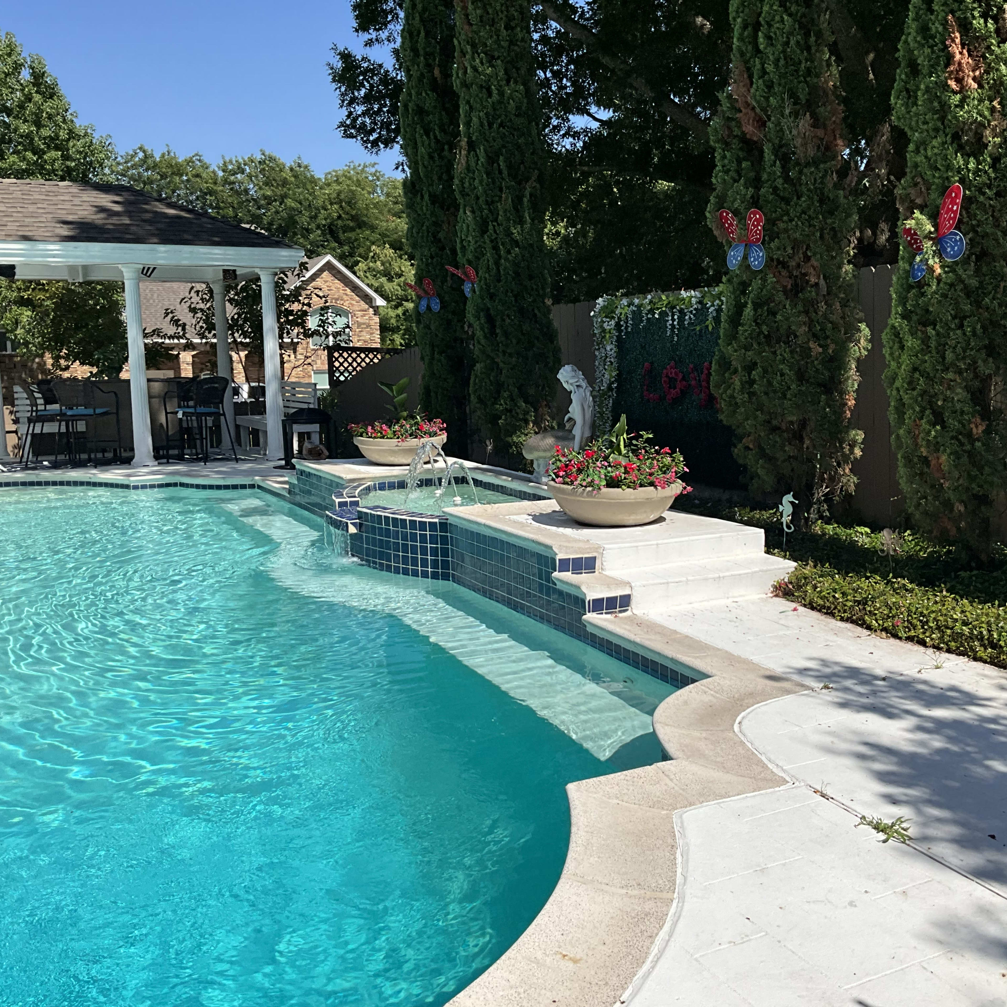 A clear swimming pool with steps leading up to a landscaped area featuring potted flowers and decorative butterflies along a fence.