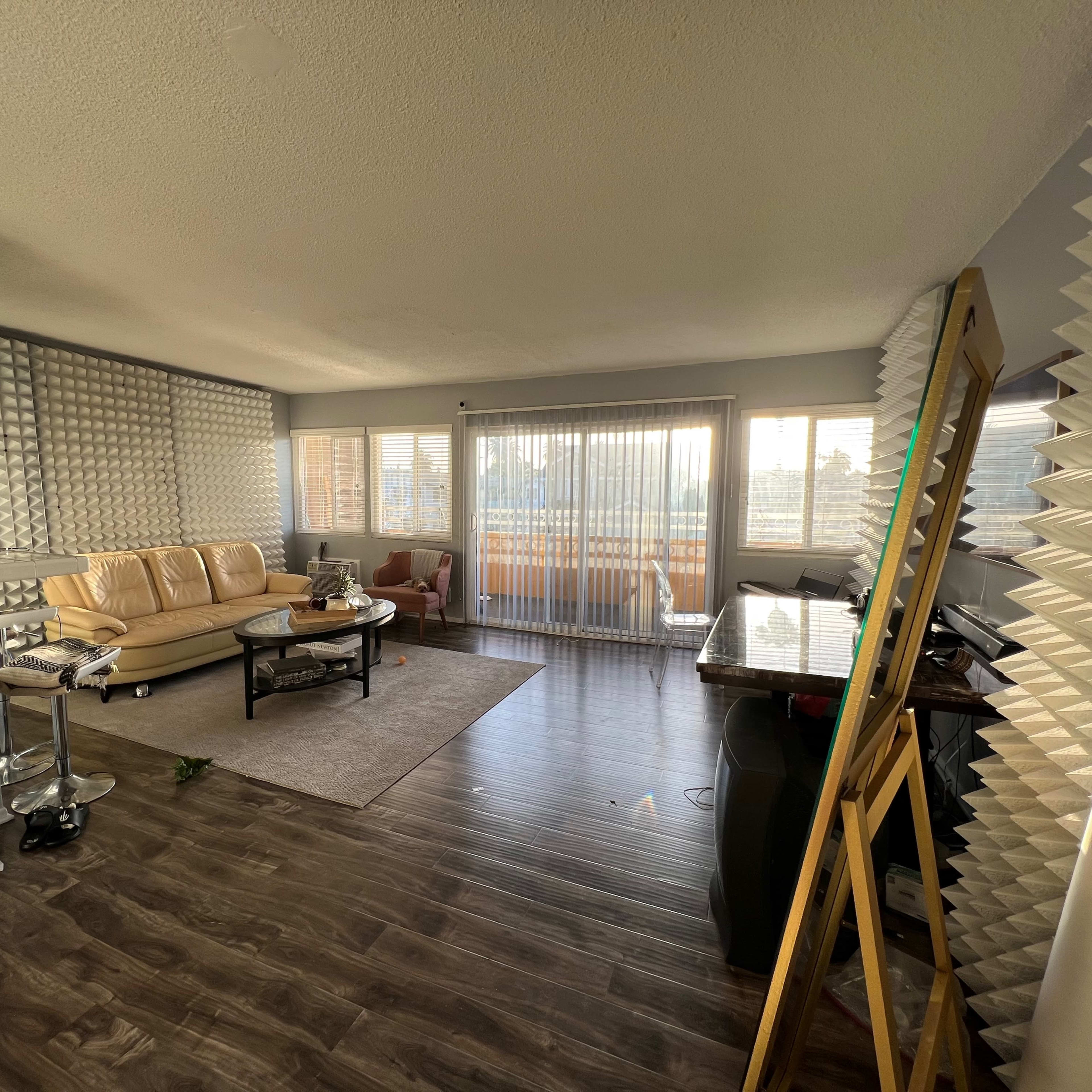 A modern living room with a beige sofa, a glass coffee table, and large windows covered by sheer curtains, set in a space with patterned wall panels.