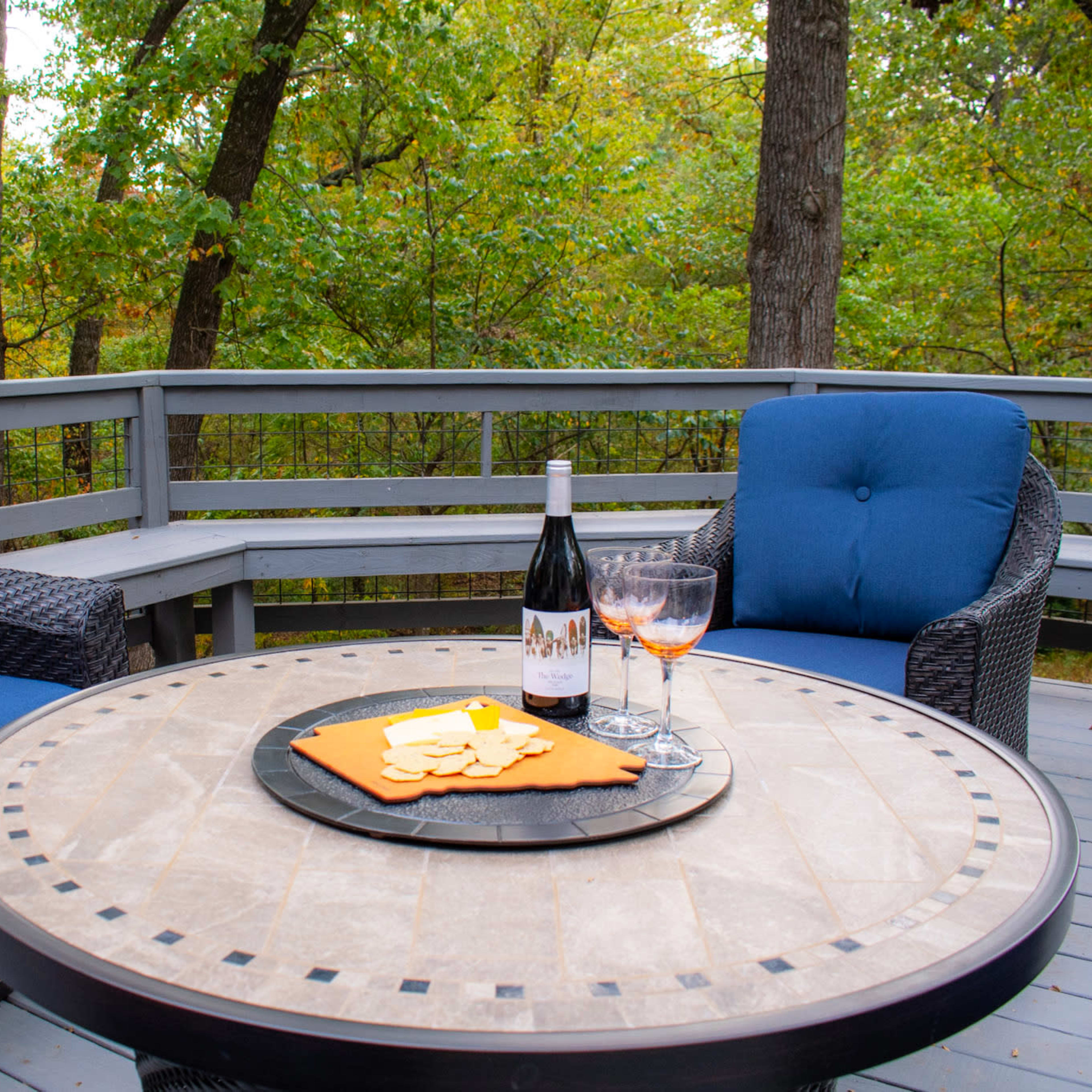 A patio setting features two blue cushioned chairs, a round table with snacks, and a bottle of wine beside two glasses against a backdrop of trees.