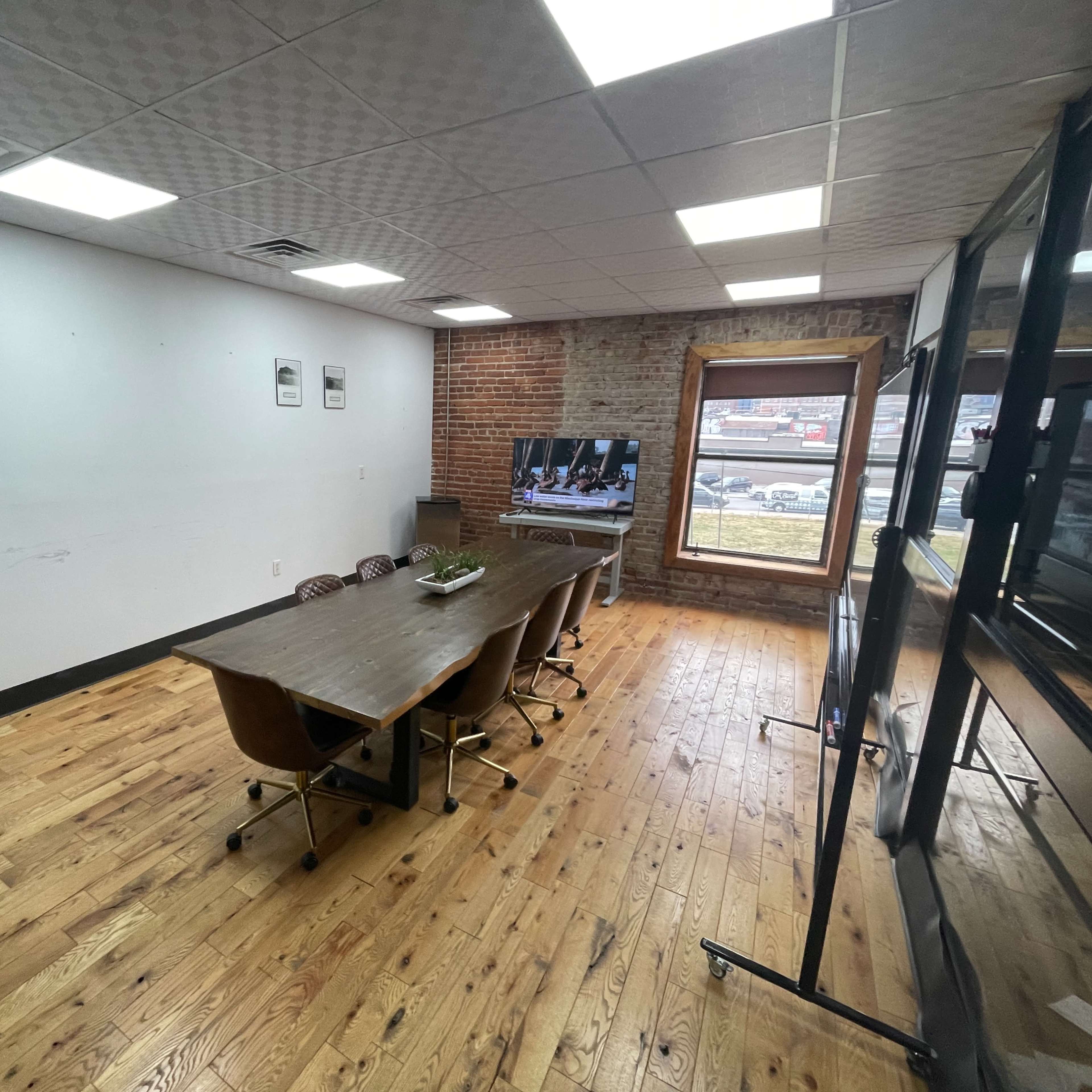 A conference room with a long wooden table surrounded by chairs, a glass-walled office section, and a large window facing the street.