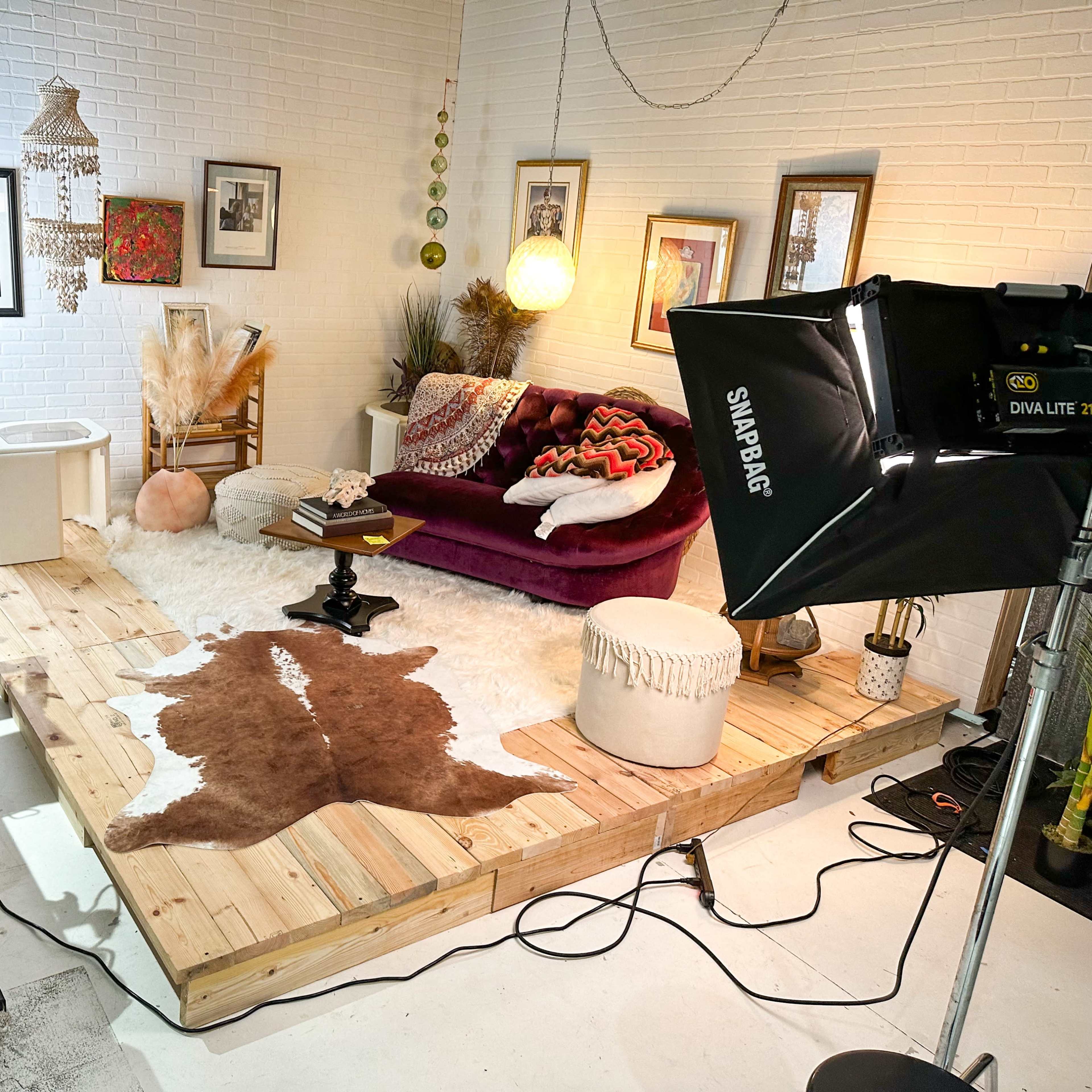 A cozy studio setup featuring a wooden platform with a cowhide rug, a burgundy sofa, and various decorative items, including a large soft light and workstations.
