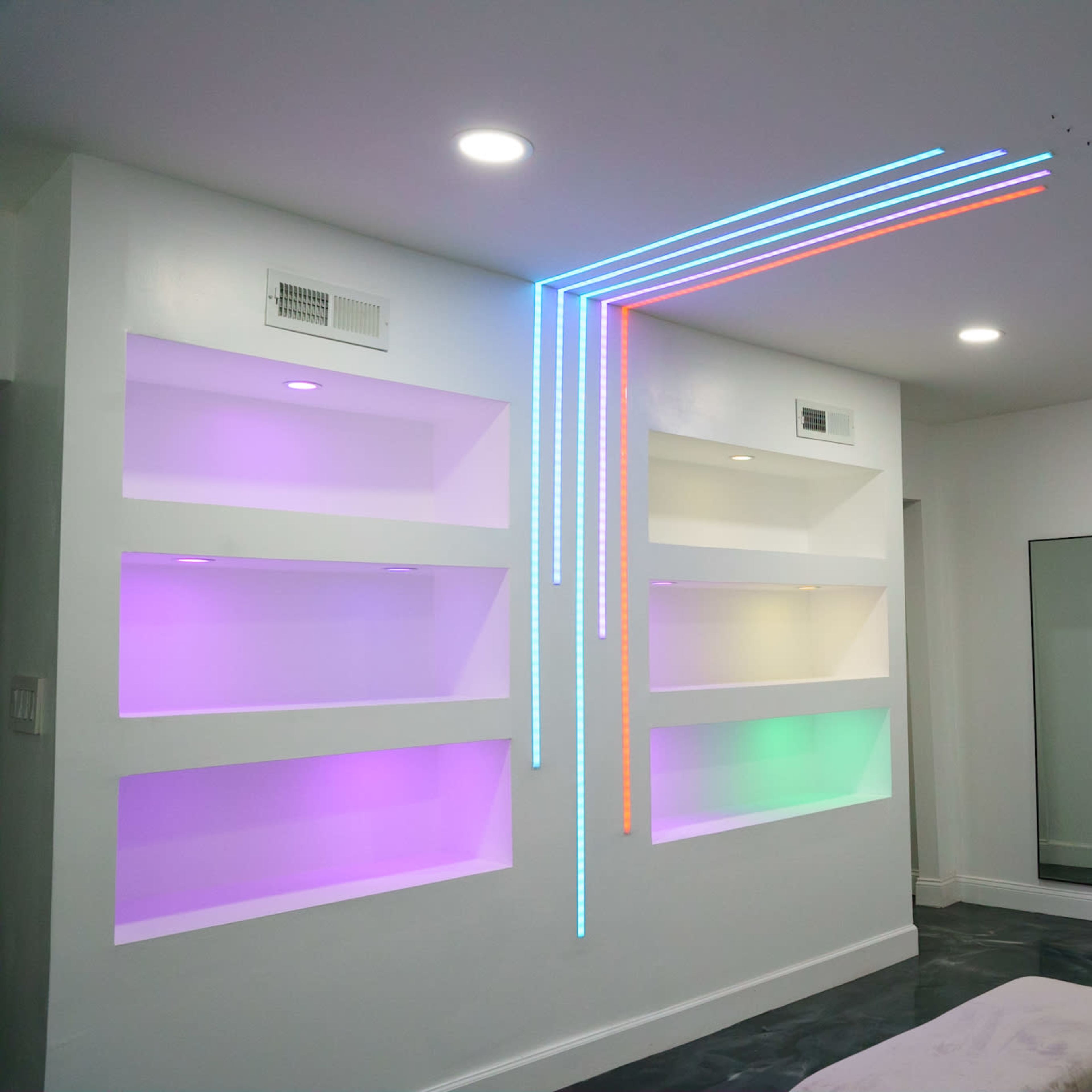 A modern living room with white walls, illuminated shelves in various colors, and linear LED lights along one wall.