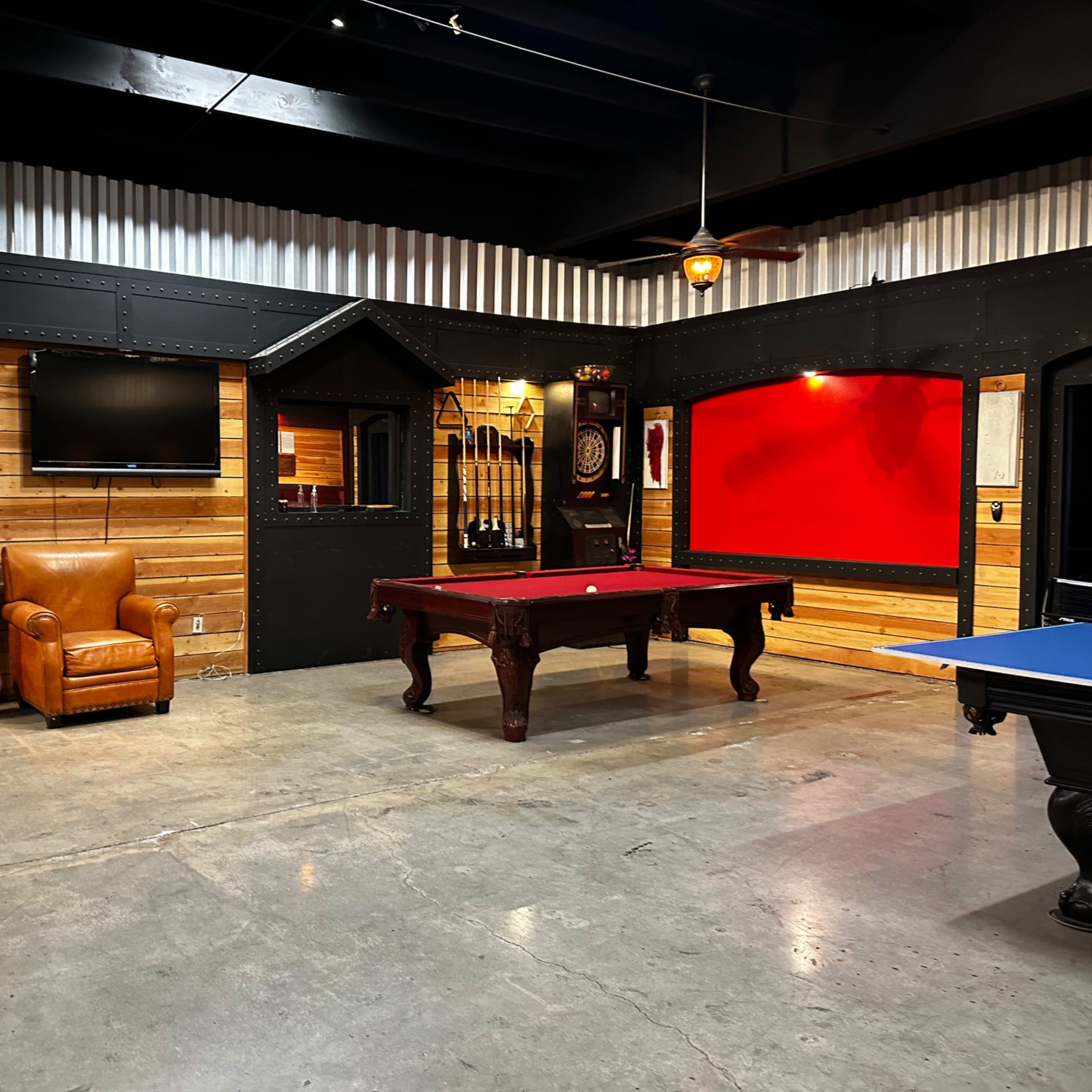 A game room featuring a pool table, a blue ping pong table, a television on the wall, and leather seating in a rustic setting with wooden and metal accents.