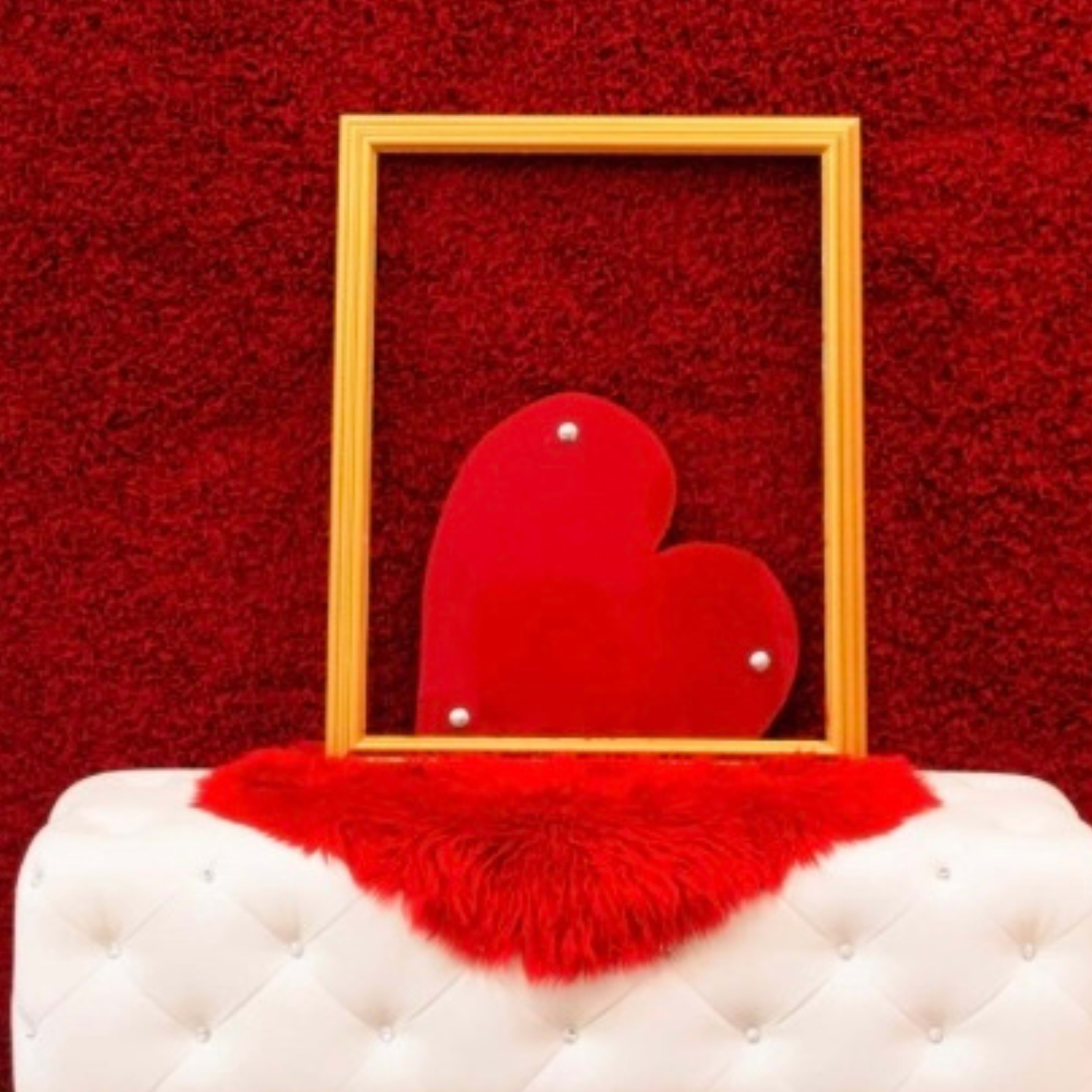 A golden picture frame displays a red heart on a white tufted bench, set against a red textured background.