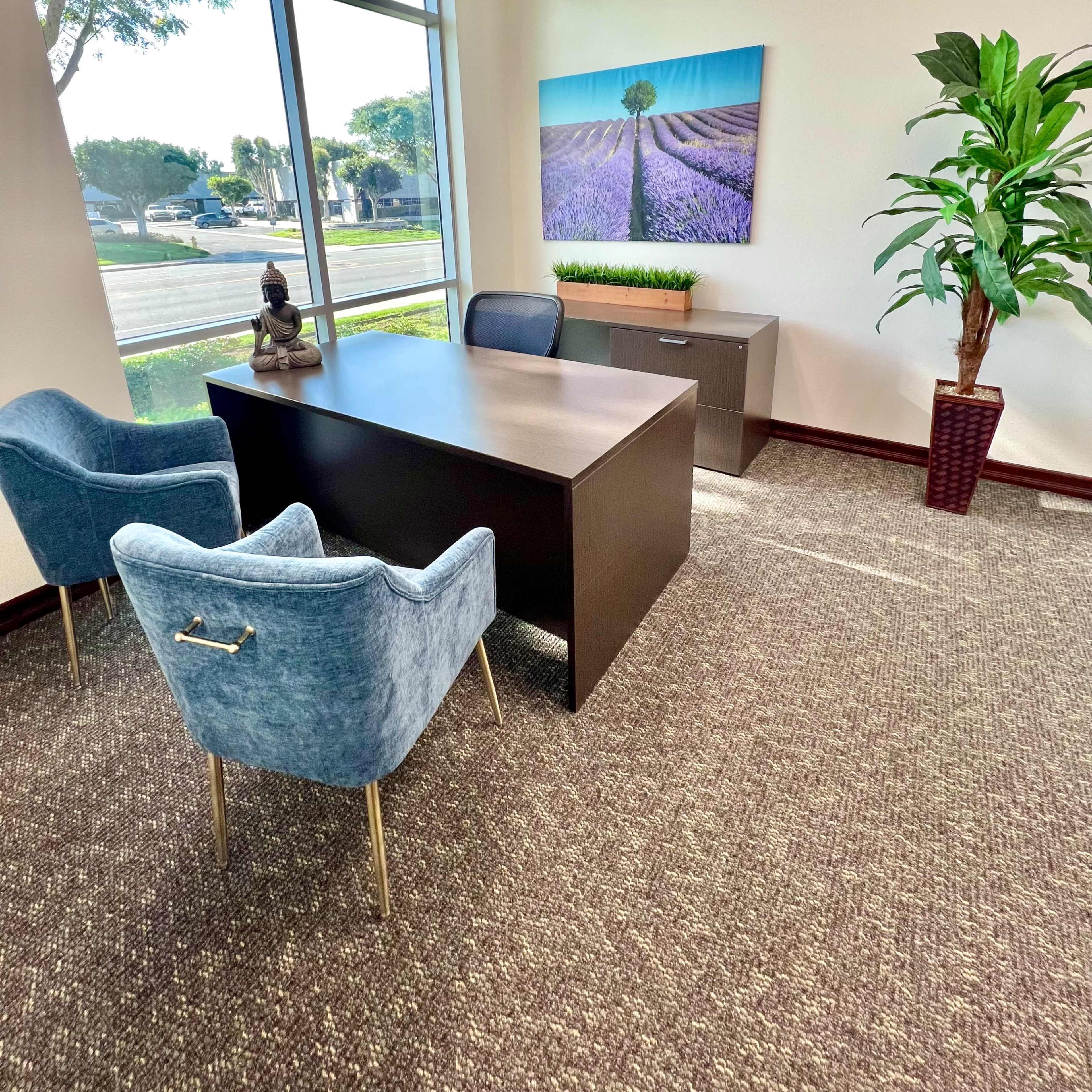 A modern office with a desk, two blue chairs, a plant, and a large wall art depicting a tree in a lavender field, all illuminated by natural light from large windows.