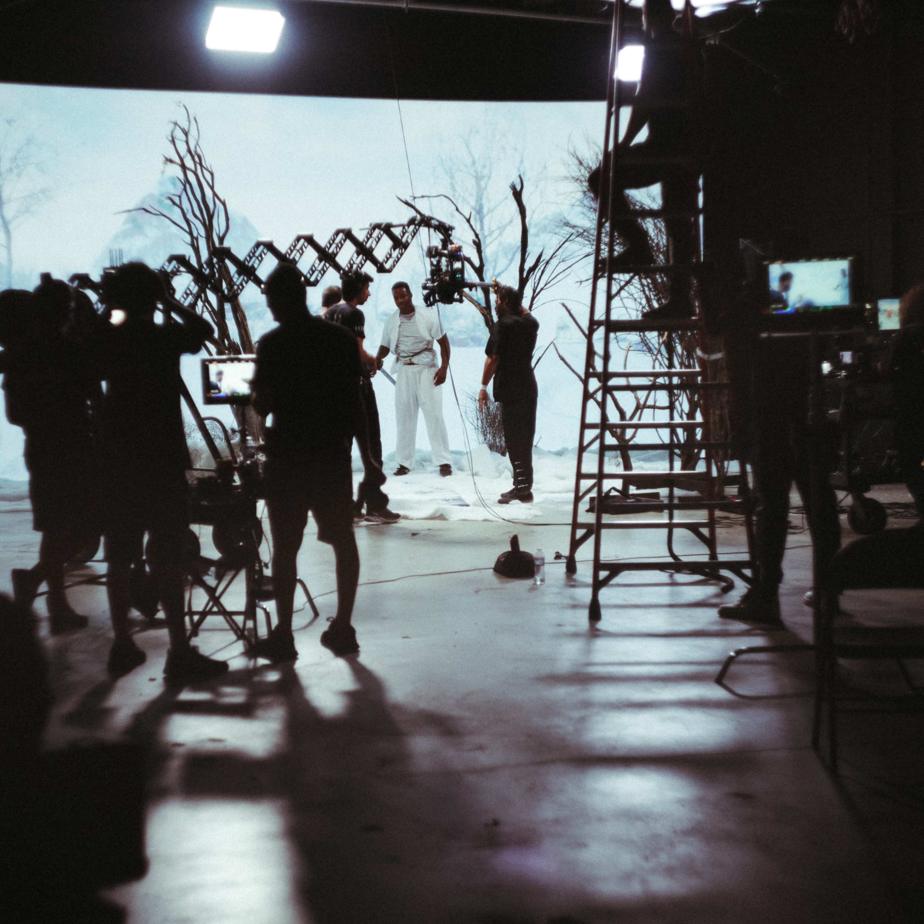 A film crew is capturing a scene in front of a white backdrop with trees, while various cameras and equipment are positioned around them.