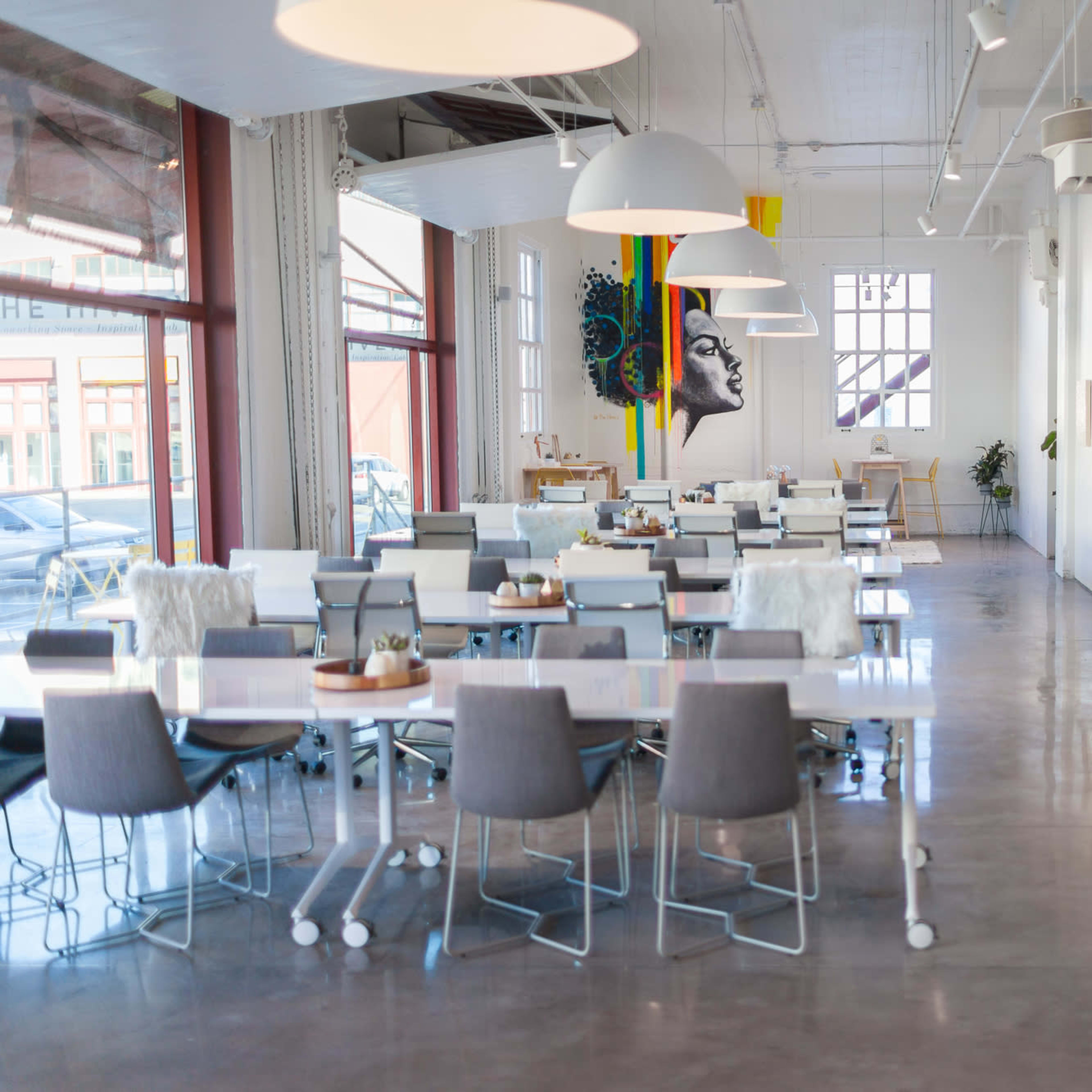 A modern café interior with white tables and chairs, large windows, and a mural on the wall.