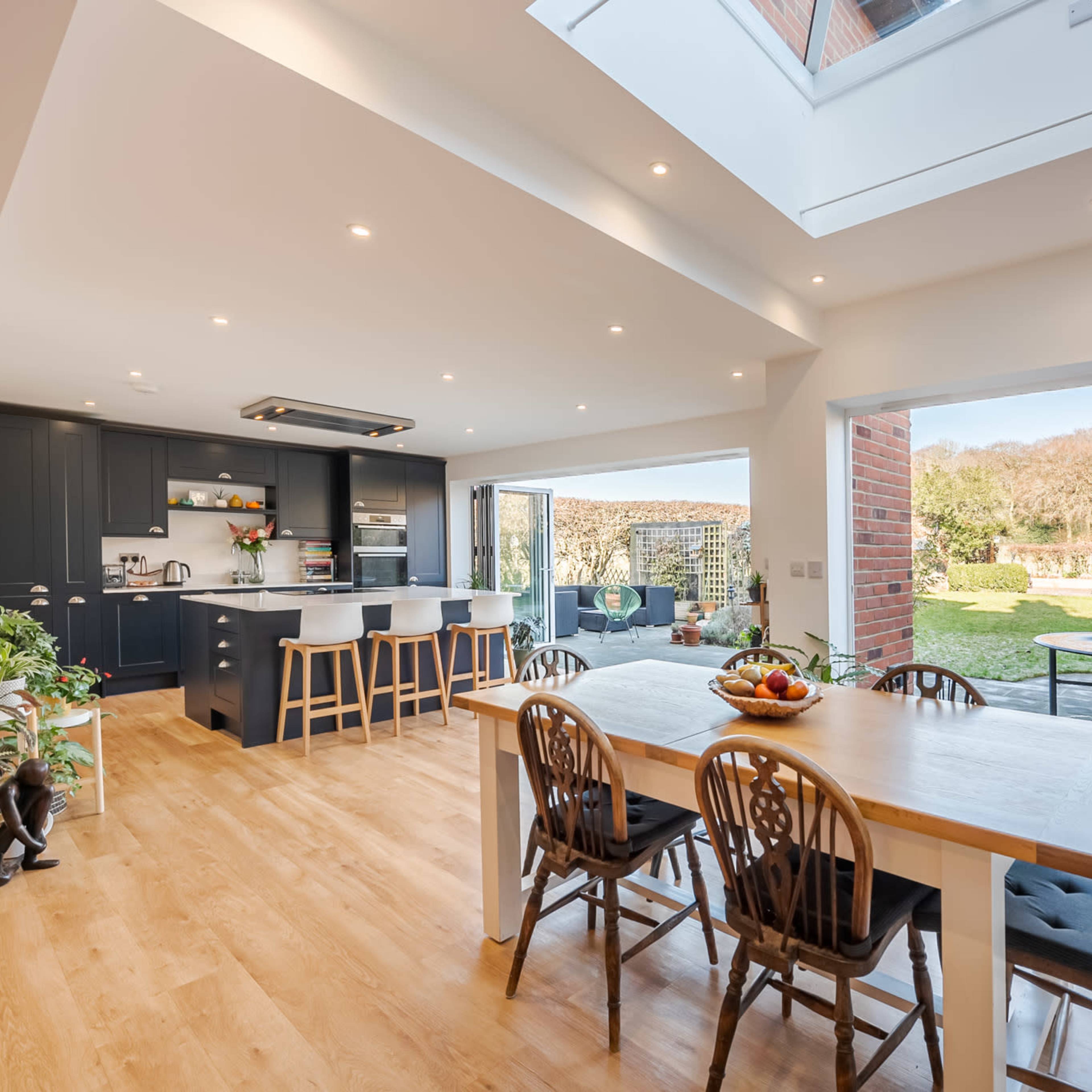 A spacious kitchen with a dining area opens to a garden through large glass doors, featuring modern furnishings and ample natural light.