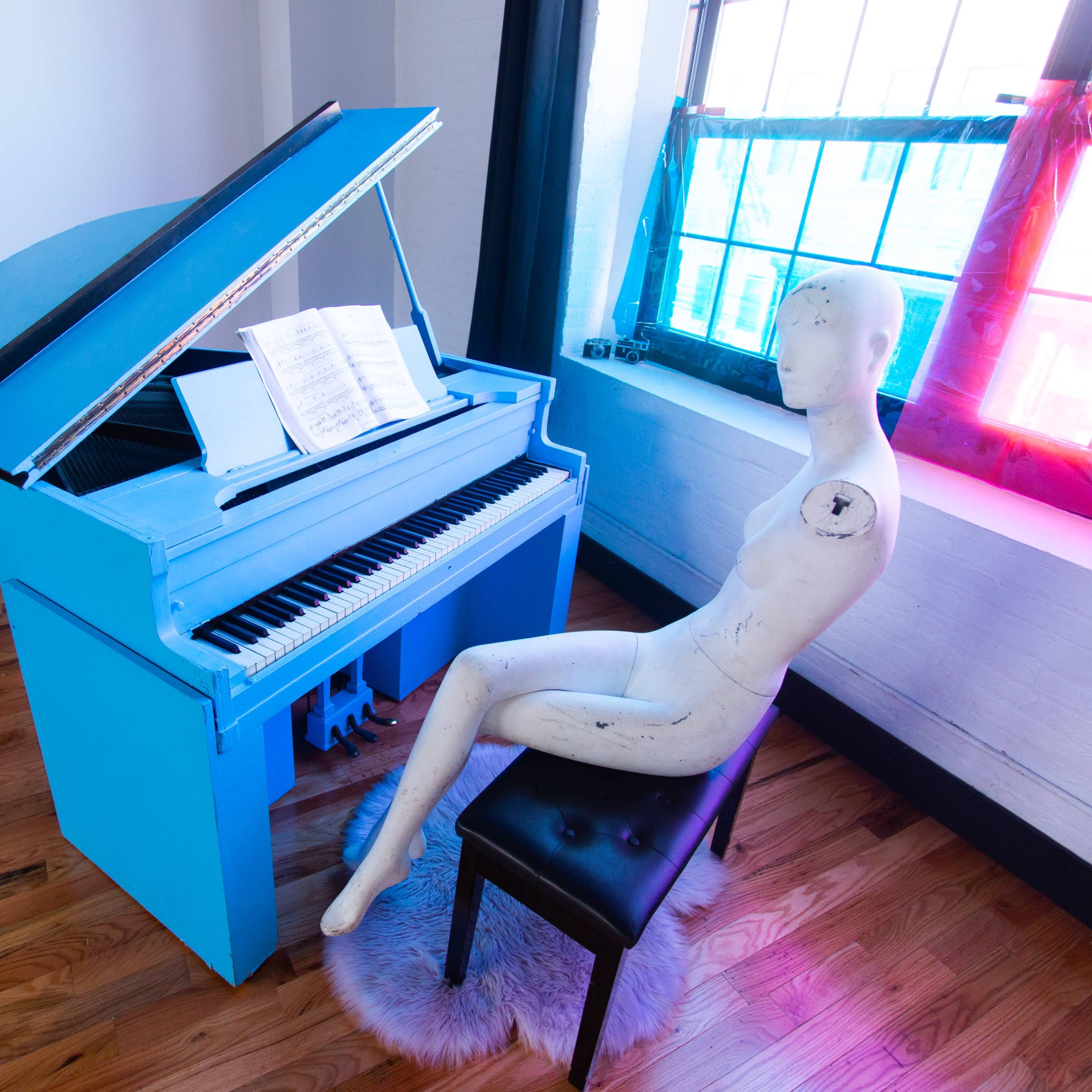 A bright blue grand piano is positioned beside a black stool with a mannequin seated on it, with sheet music displayed on the piano.