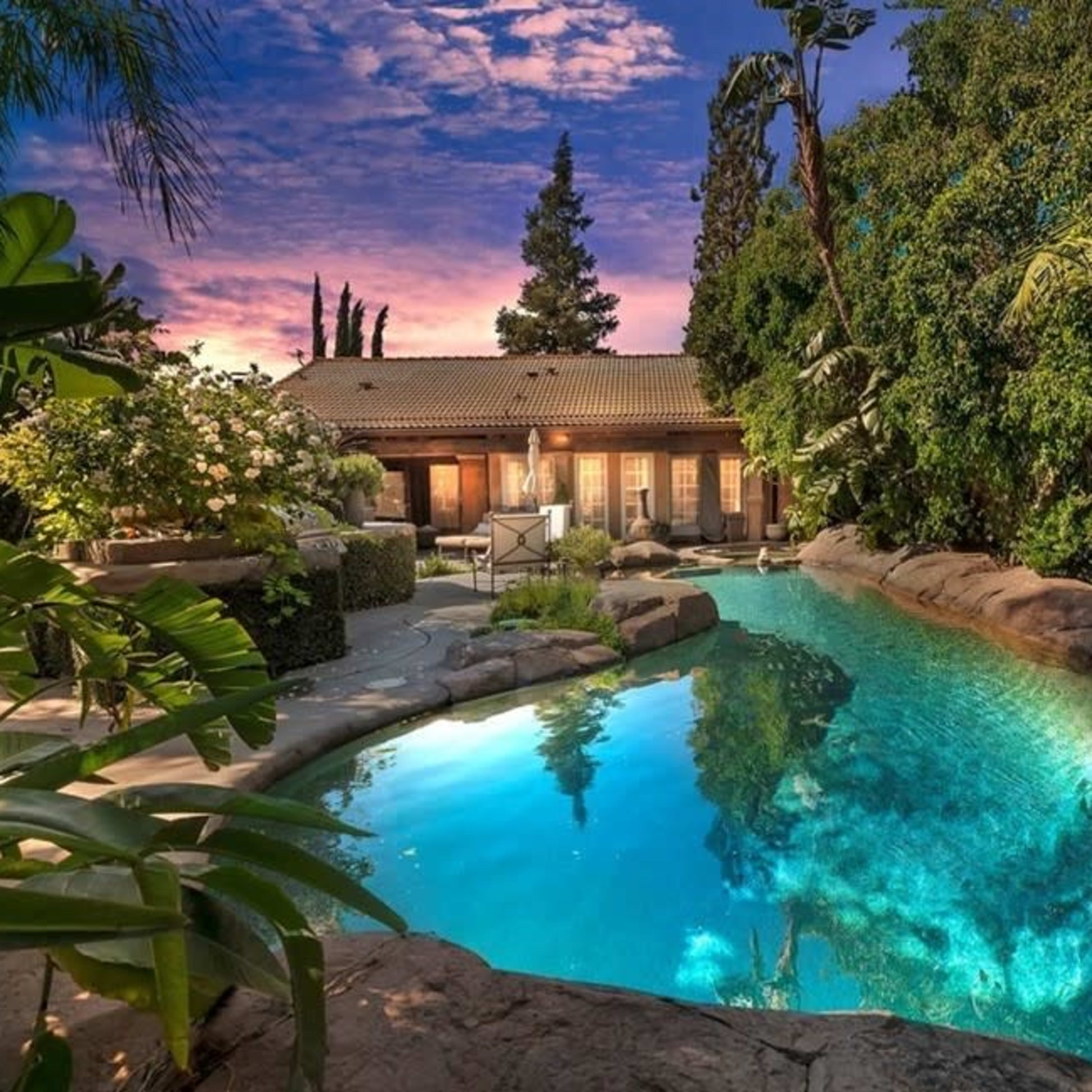 A resort-style swimming pool surrounded by lush greenery and a backdrop of a house at sunset.