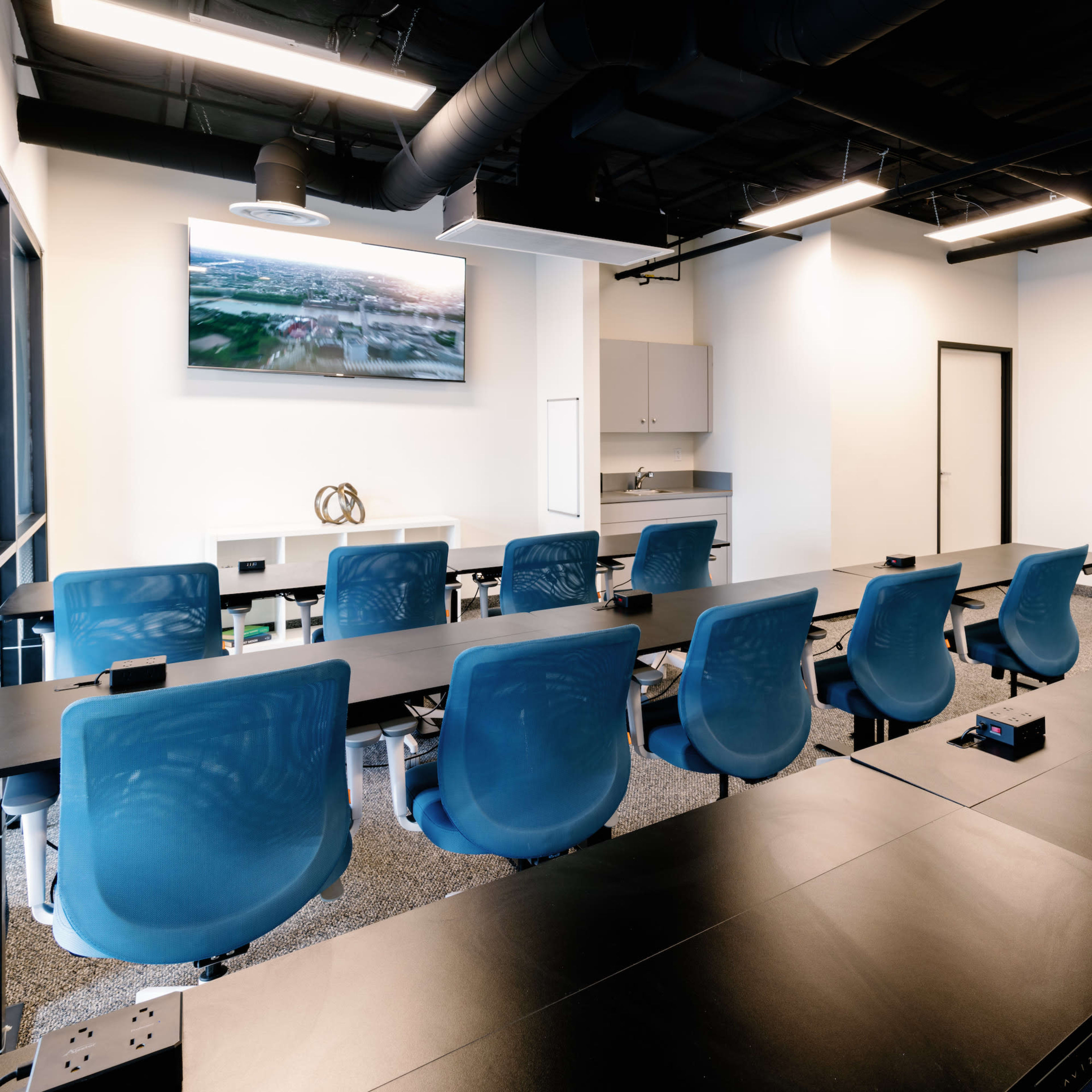 A modern conference room features a long black table with blue chairs, a television mounted on the wall, and a kitchenette area.