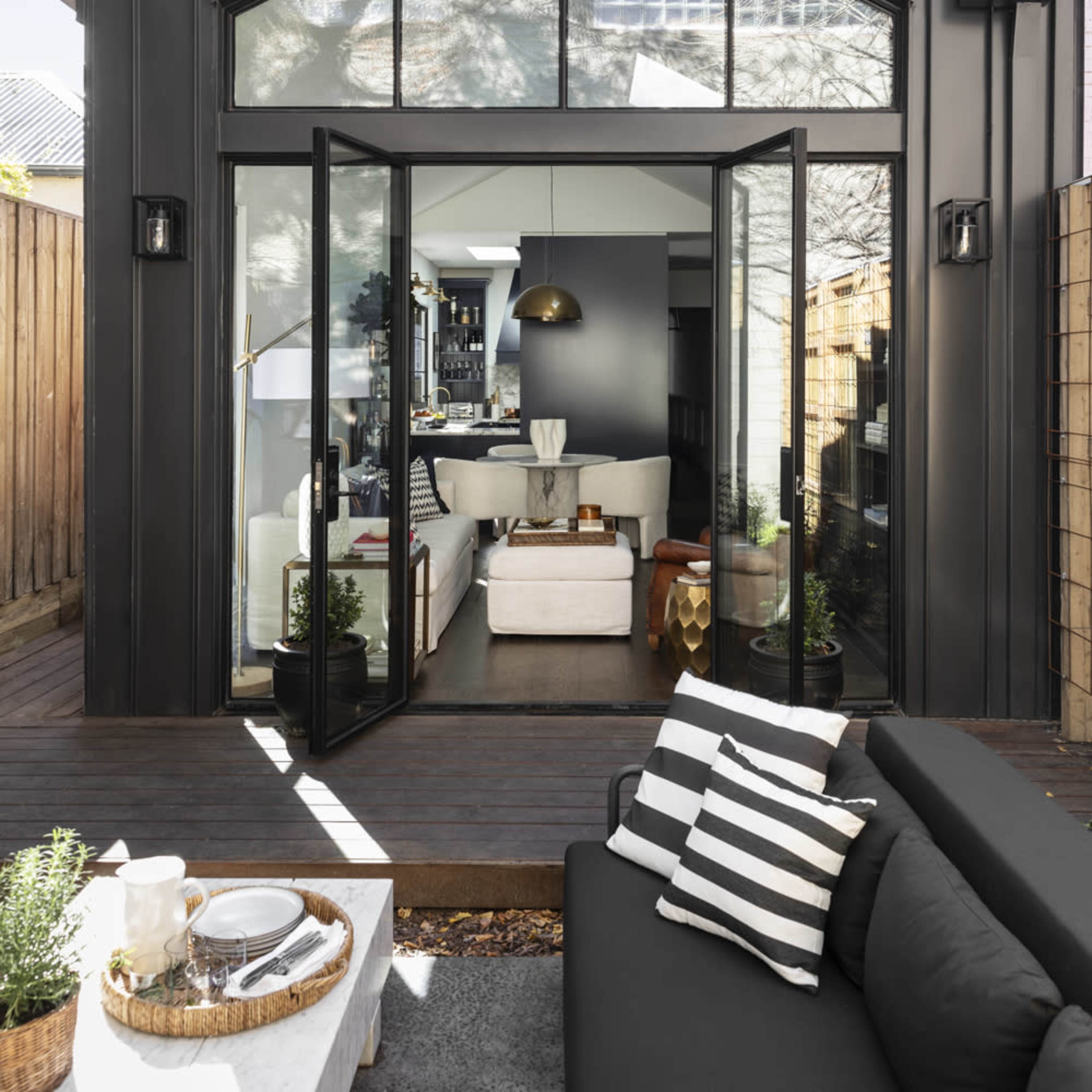 A modern black house with large glass doors opening to a living space, surrounded by a wooden deck and outdoor seating.