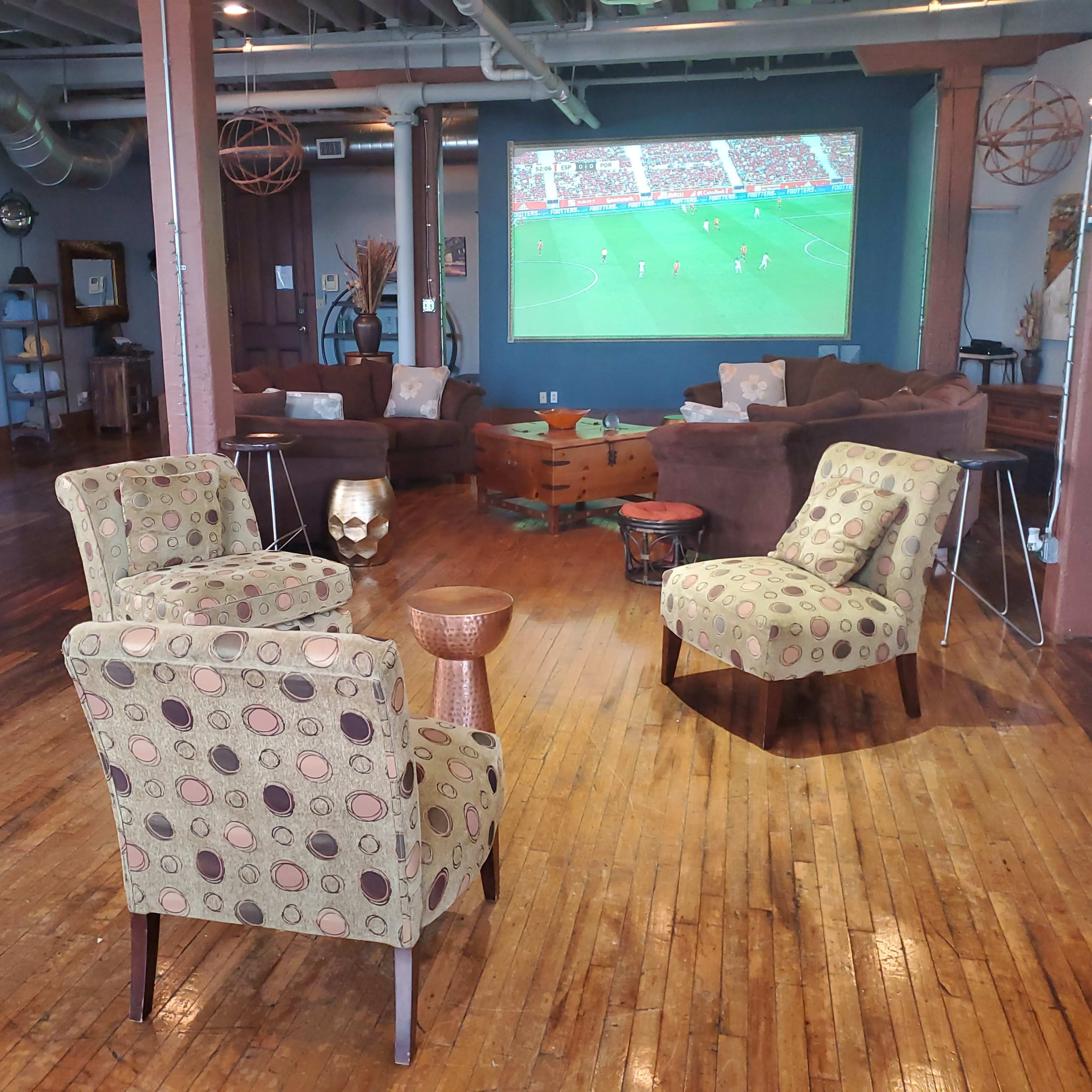 A spacious room with wooden floors, featuring three patterned chairs and a small table, while a large screen displays a soccer match in the background.