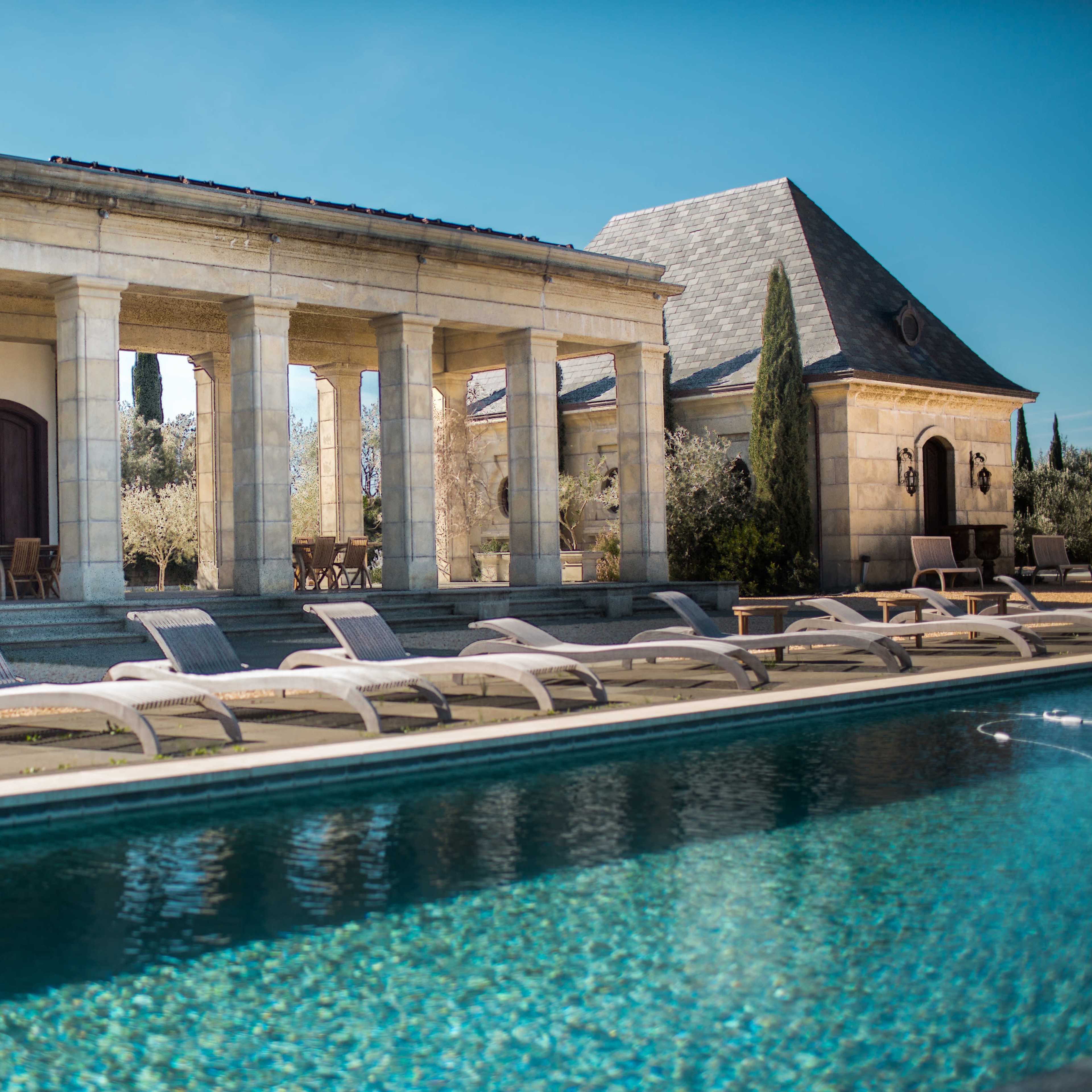 A serene outdoor swimming pool lined with lounge chairs in front of a stone building with columns and a roofed area.