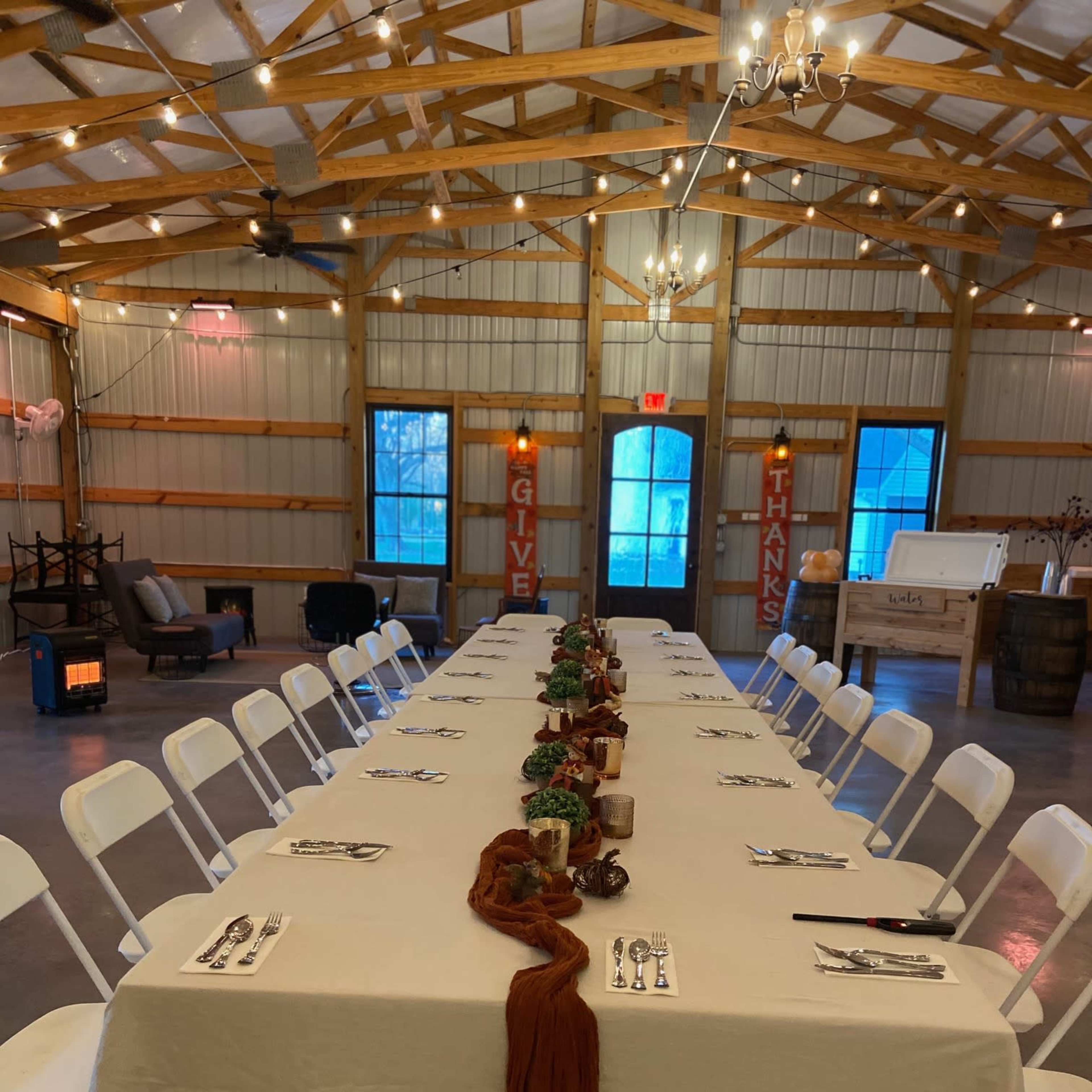 A long, neatly arranged dining table with white chairs is set up in a spacious hall featuring wooden beams, string lights, and decorations that express gratitude.